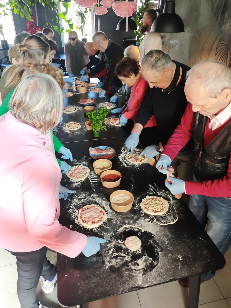 Zdjęcie przedstawia grupę seniorów przy stole wykonujących samodzielnie pizze z dostępnych składkników