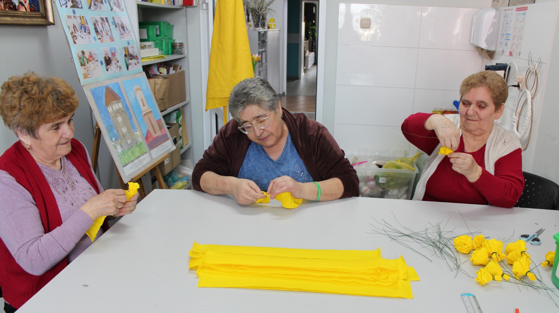 Zdjęcie wykonane w pracowni plastycznej. Przedstawia trzy seniorki robiące kwiaty w krepiny