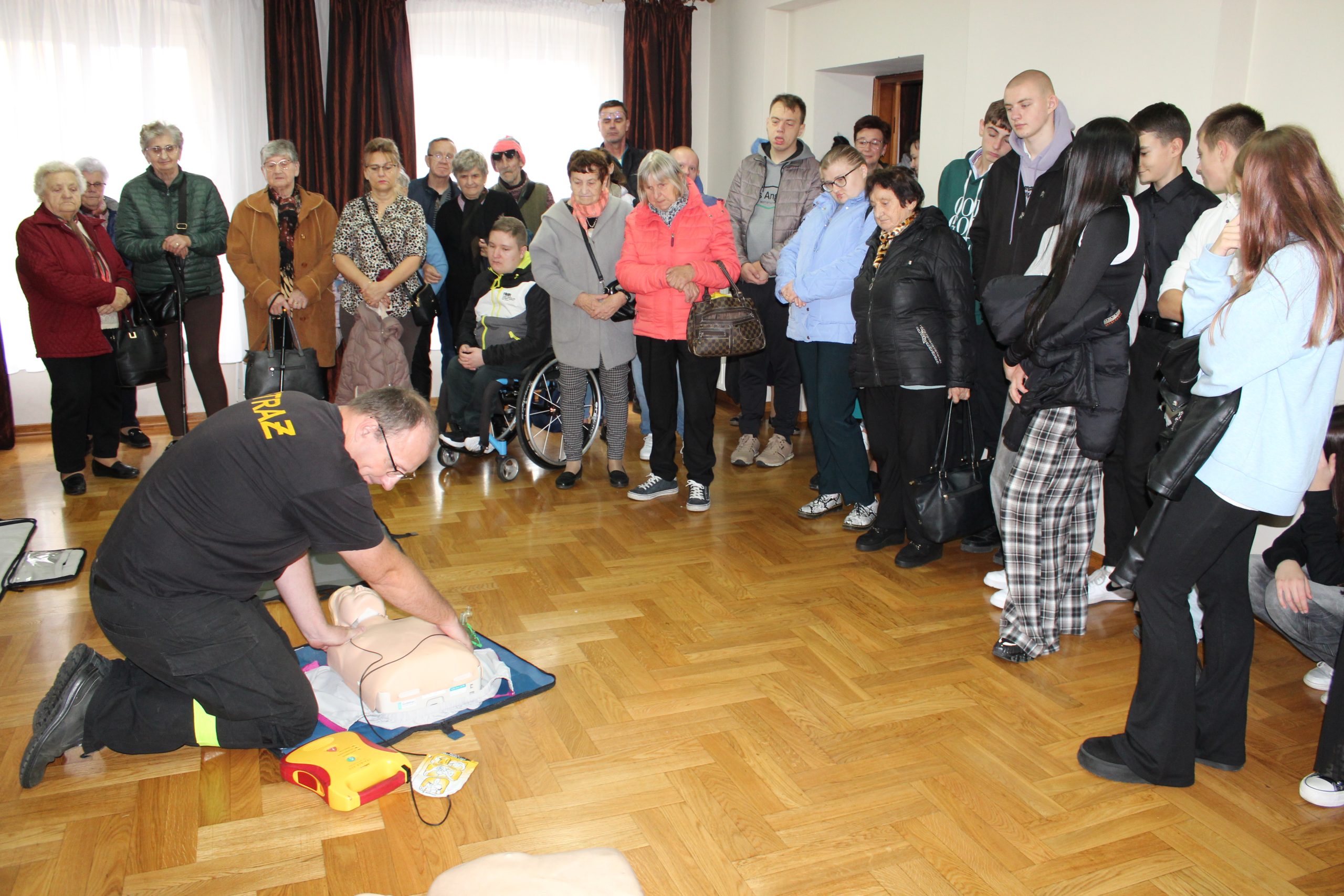 3) Zdjęcie wykonane w sali MGOK w Sompolnie. Przedstawia młodzież oraz seniorów zgromadzonych podczas pokazu Pierwszej Pomocy z użyciem defibrylatora (AED)