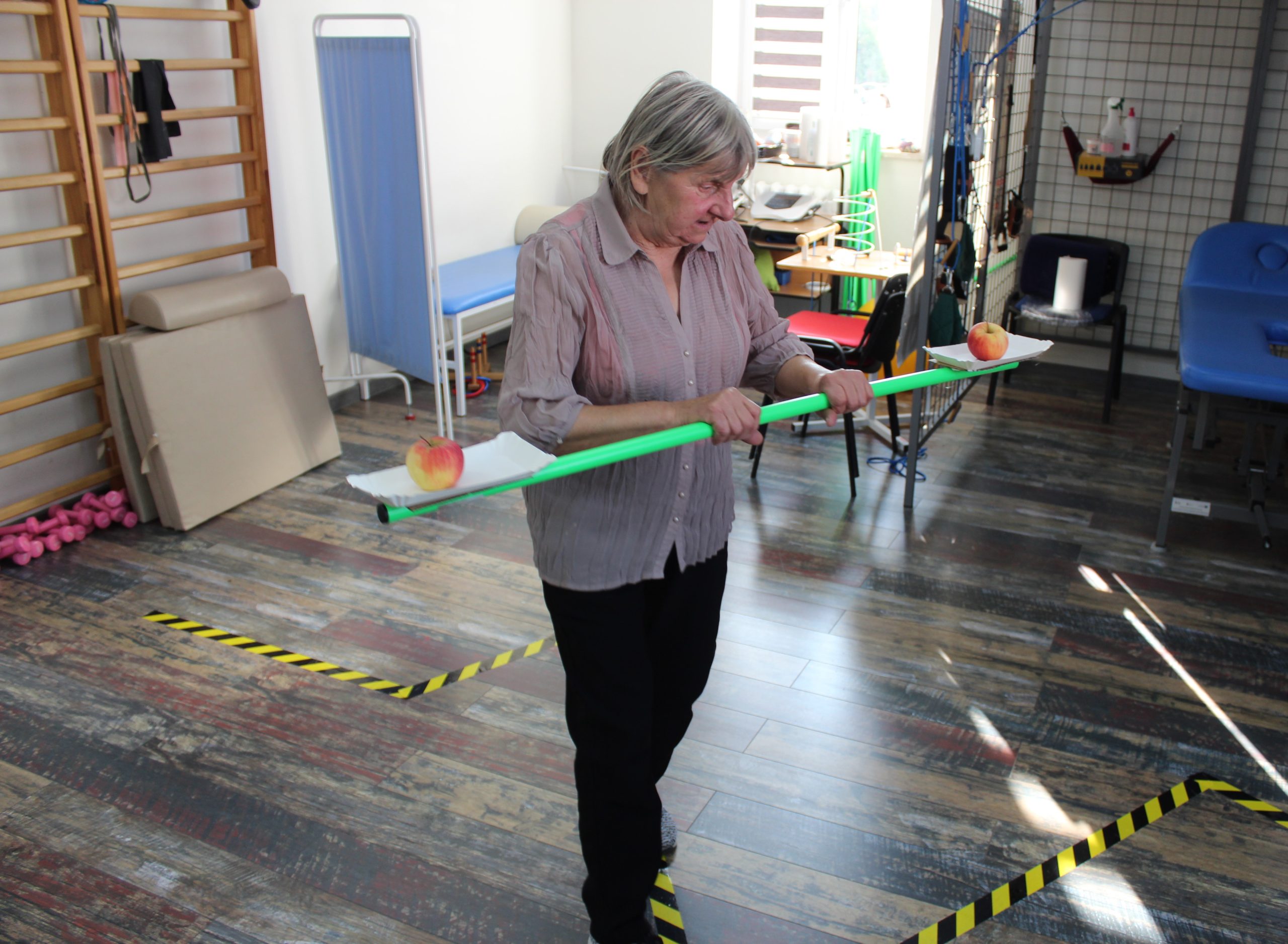 3) Zdjęcie wykonane na sali rehabilitacyjnej w DDP. Przedstawia Seniorkę pokonującą slalom tak by nie upuścić trzymanych na tackach jabłek.