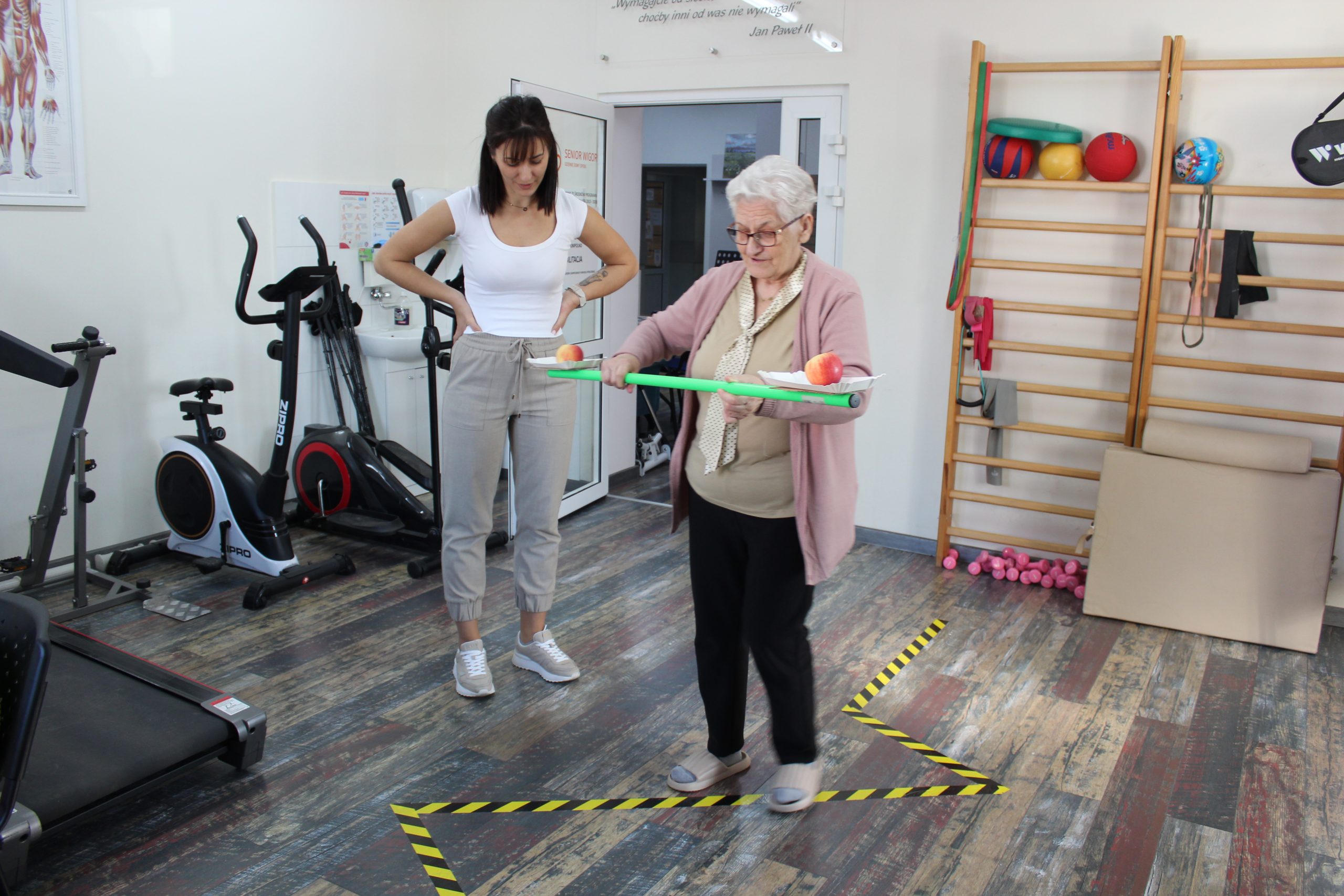 4) Fotografia przedstawia seniorkę podczas konkurencji ruchowej wykonywanej na sali rehabilitacyjnej DDP.