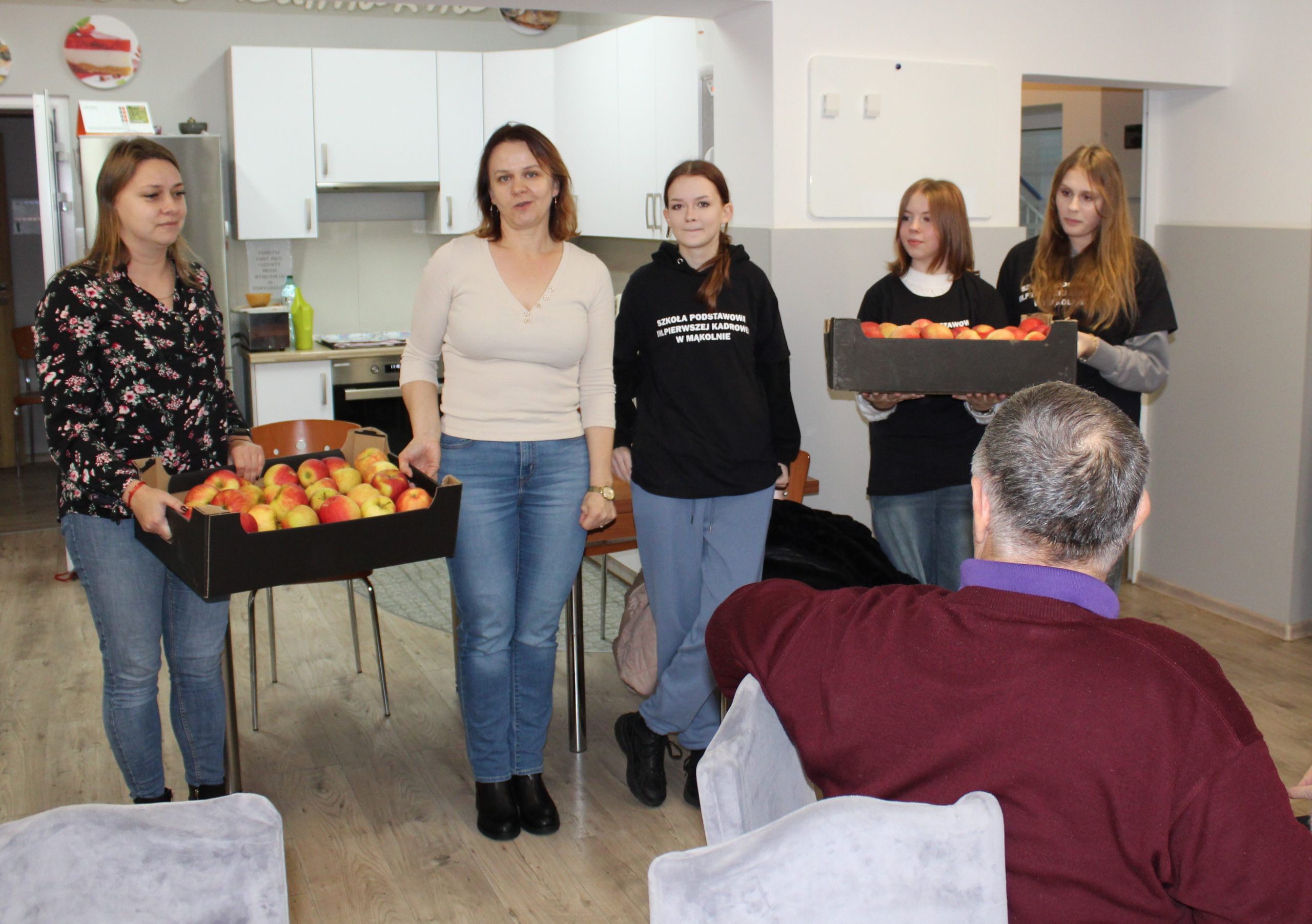 1. Zdjęcie wykonane na sali głównej Domu Dziennego Pobytu. Przedstawia Wolontariuszy wraz z opiekunami wręczających Seniorom jabłka.