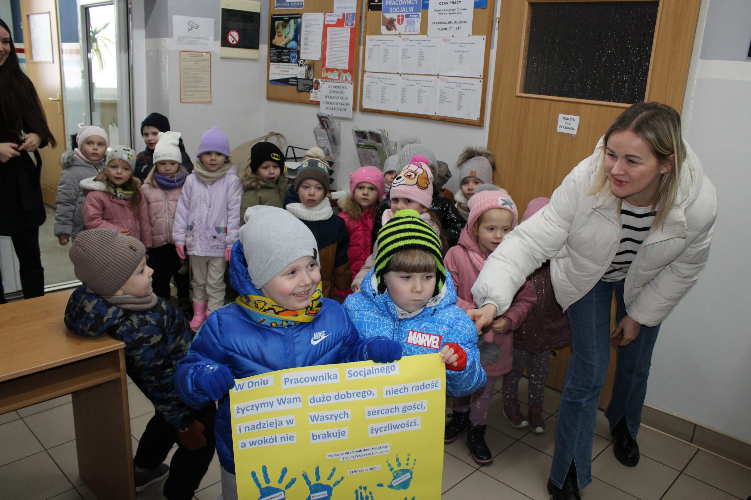 Zdjęcie wykonane w Ośrodku Pomocy Społecznej w Sompolnie. Przedstawia przedszkolaków trzymających dużą laurkę wykonaną z okazji dnia pracownika socjalnego oraz panie nauczycielki.