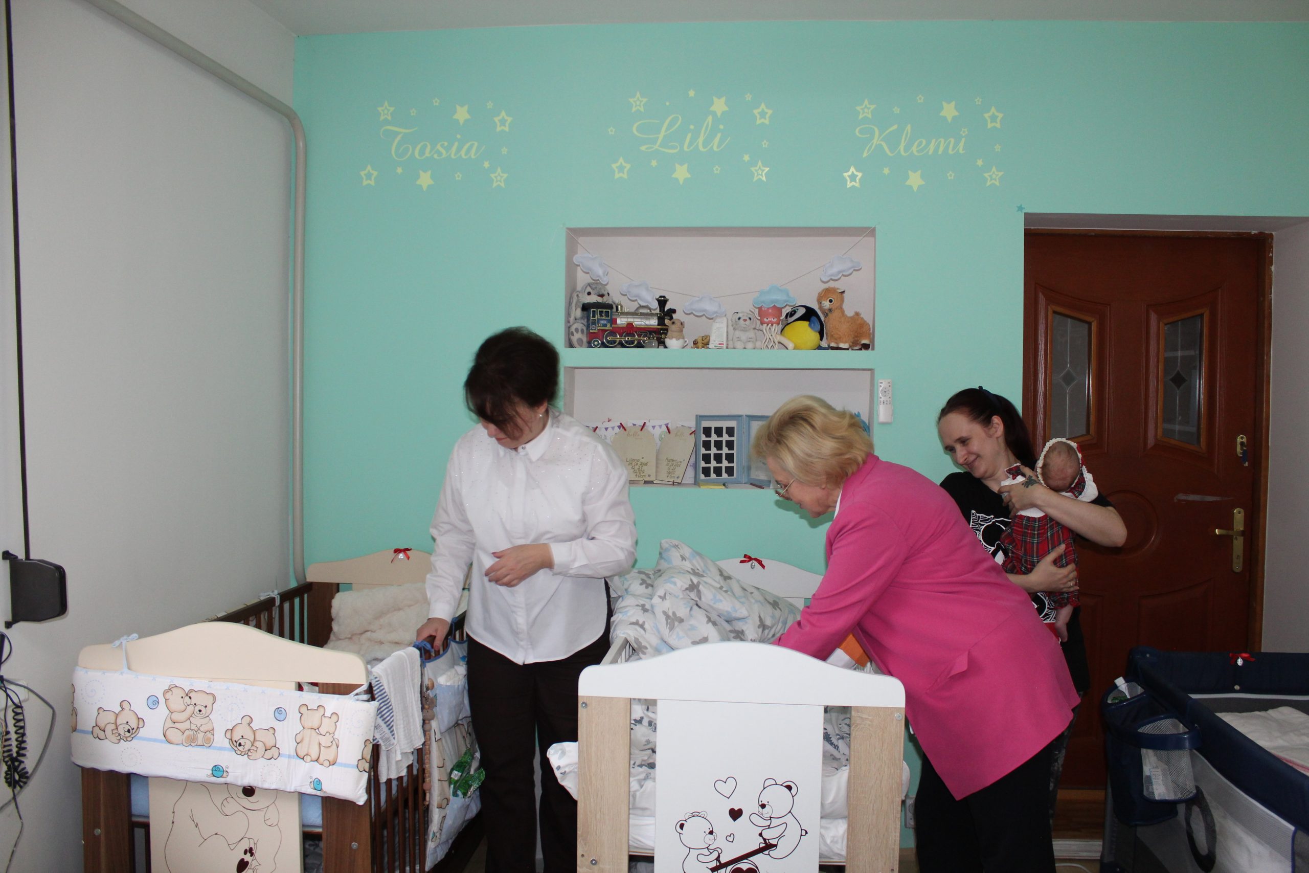 7) Na zdjęciu trzy kobiety stojące przy łóżeczkach dzieci