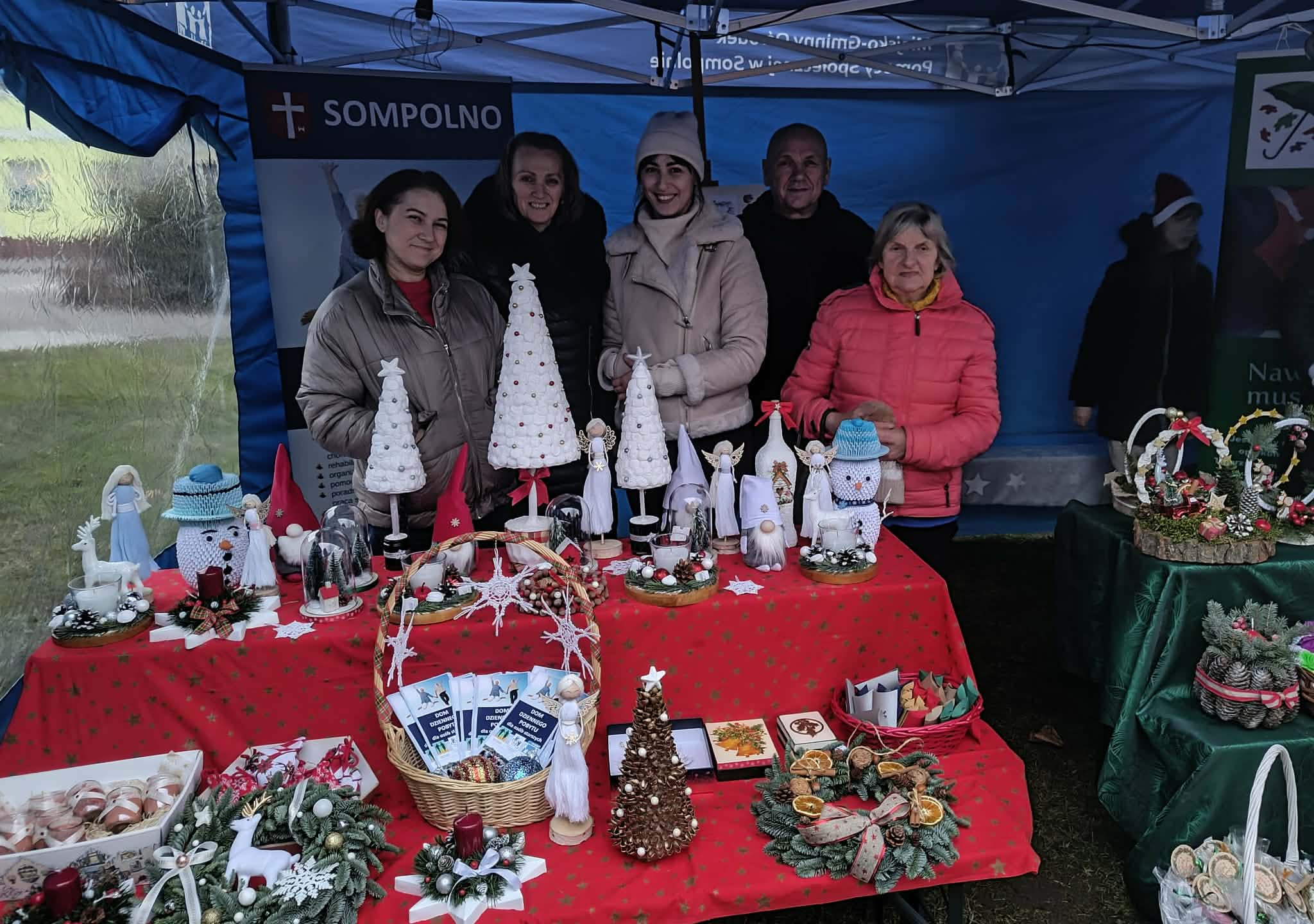 1) Zdjęcie wykonane podczas kiermaszu bożonarodzeniowego. Przedstawia pracowników przy stoisku DDP. Przed nimi stół z rękodziełem.