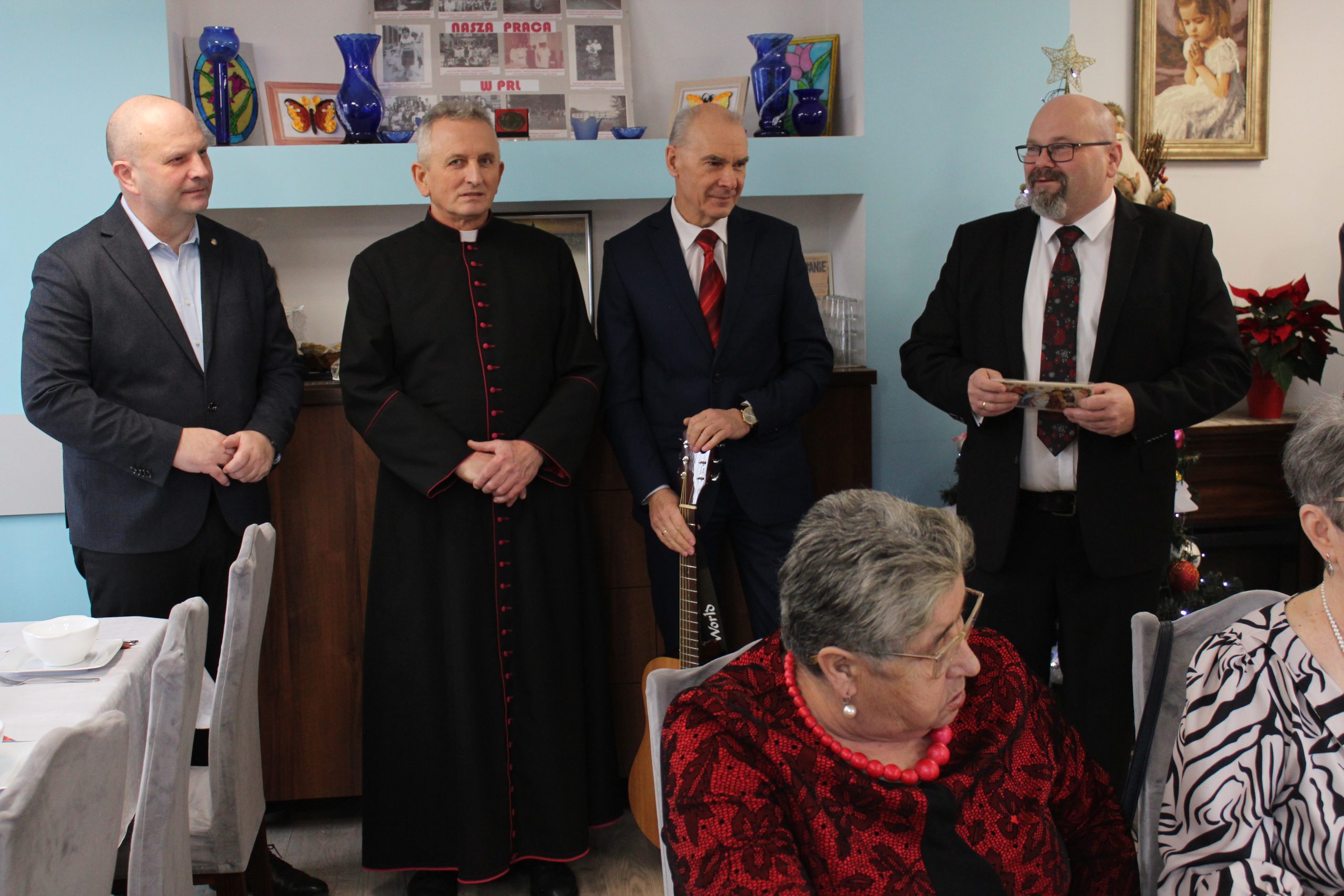 1) Zdjęcie wykonane w sali głównej Domu Dziennego Pobytu. Przedstawia od lewej: burmistrza, księdza, pracownika ddp stojącego z gitarą oraz dyrektora mgops.