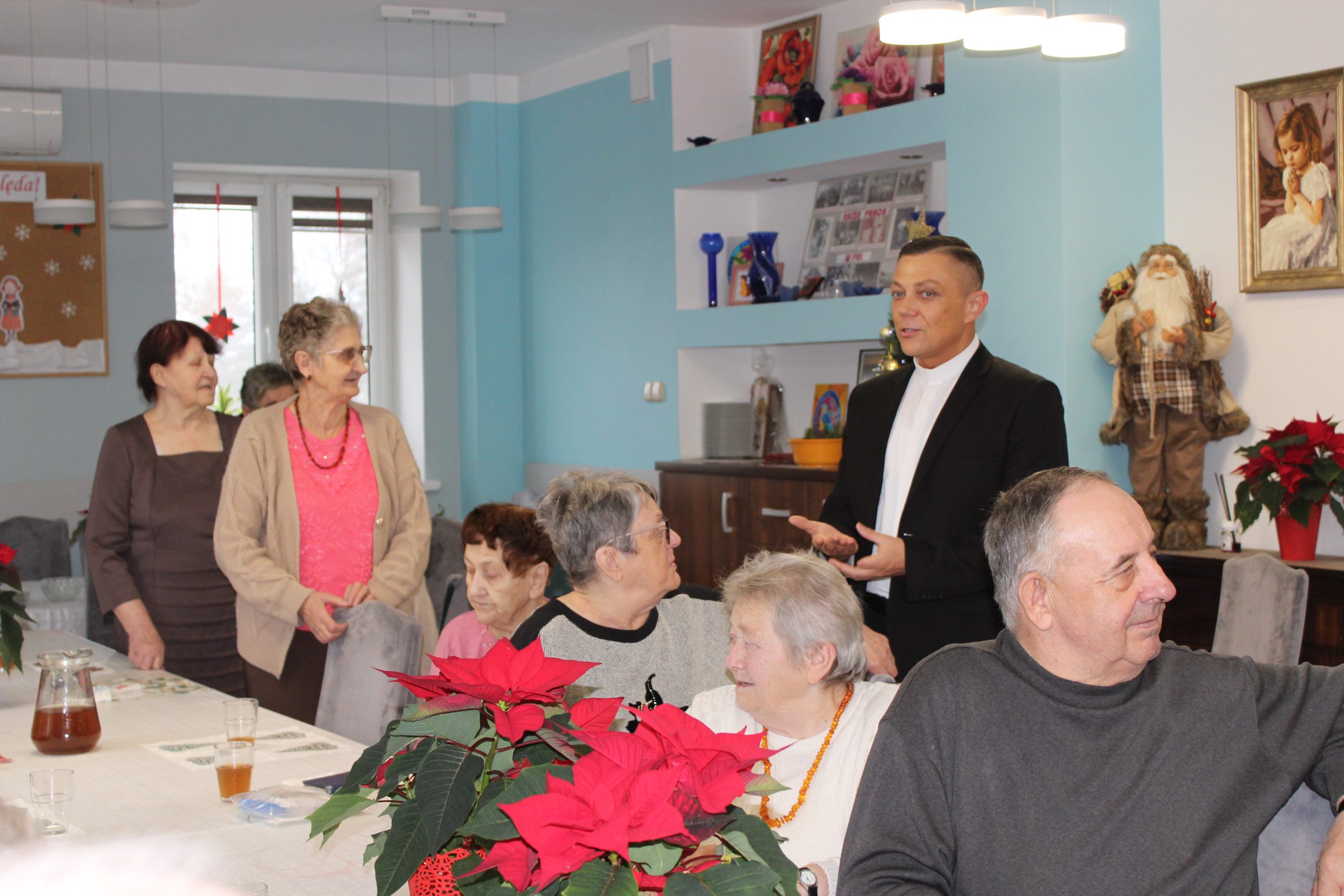 1) Zdjęcie wykonane w Domu Dziennego Pobytu. Przedstawia seniorów siedzących przy stole oraz księdza rozmawiającego z nimi.