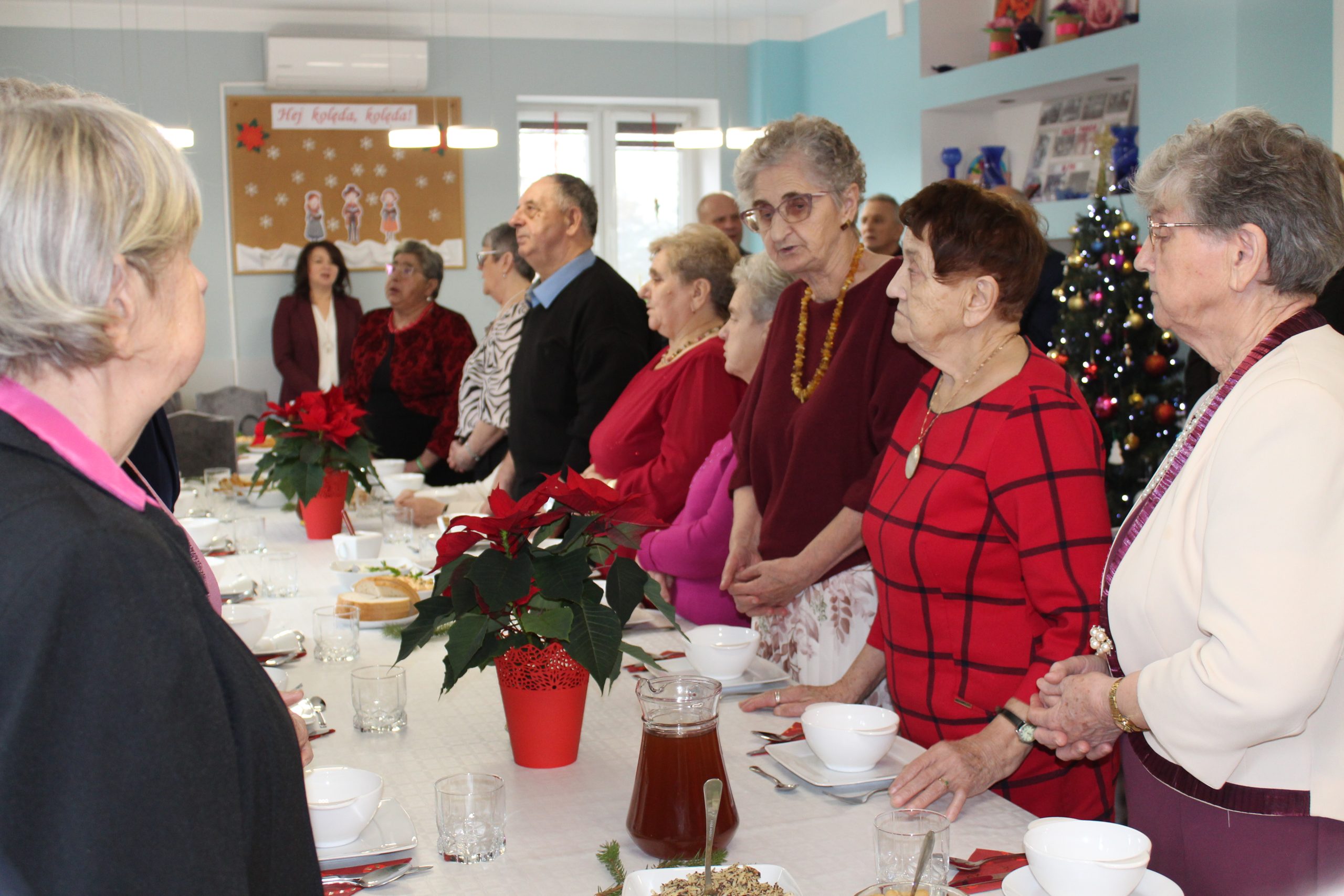 3) Na zdjęciu seniorzy stojący przy stołach i śpiewający kolędę.