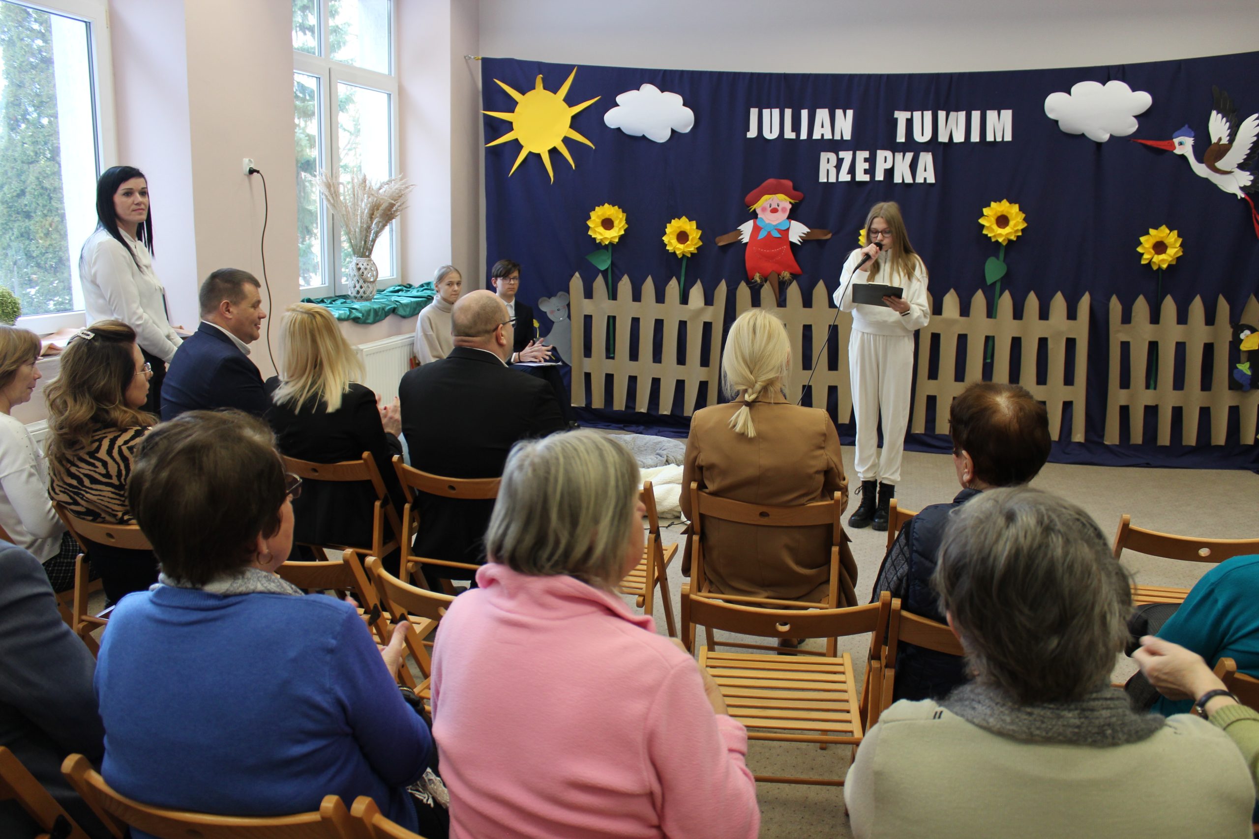 1) Zdjęcie wykonane w jednej z sal szkoły w Mąkolnie. Z tyłu granatowa gazetka rozmieszczona na całej ścianie. Na niej płot, strach na wróble, słoneczniki i napis Julian Tuwim „Rzepka”. Na tle gazetki dziewczyna z mikrofonem. Z przodu widownia siedząca na krzesłach.