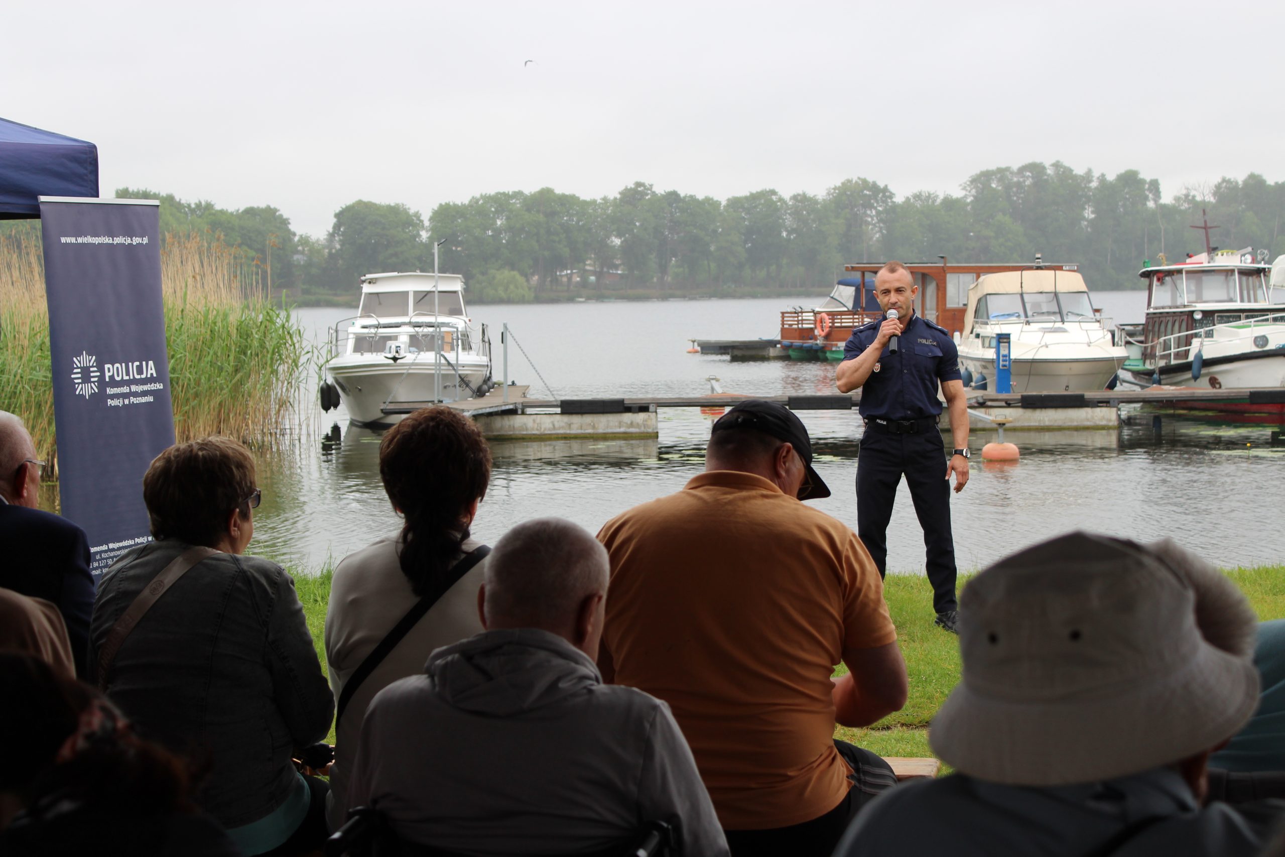 1. Zdjęcie przedstawia policjanta stojącego nad jeziorem ślesińskim, który przemawia do grupy seniorów.