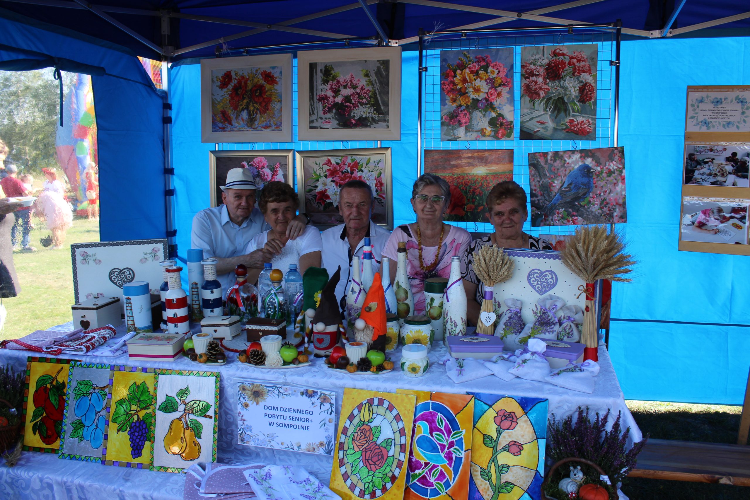 1) Zdjęcie wykonane pod namiotem domu dziennego pobytu podczas dożynek powiatowych w Lubstowie. Przedstawia grupę uczestników DDP. W tle obrazy, a z przodu wystawa rękodzieła