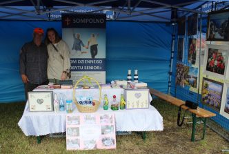 1. Zdjęcie przedstawia stoisko Domu Dziennego Pobytu dla Osób Starszych w Sompolnie podczas XXV Wielkopolskiego Pikniku Sołtysów. Na stosiku znajduje się rękodzieło oraz zdjęcia Seniorów podczas ich pracy. Za stosikiem stoi uczestnik oraz pracownica DDP