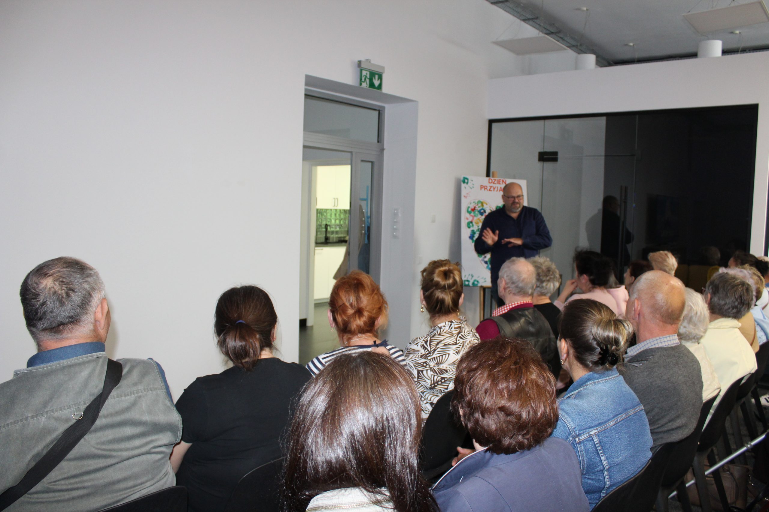 1) Zdjęcie wykonane w Dziennym Centrum Wsparcia w Sompolnie. Przedstawia Pana Tomasza Majewskiego – dyrektora M-GOPS w Sompolnie opowiadającego o nowo powstałej placówce. Przed nim uczestnicy spotkania siedzący na krzesłach.
