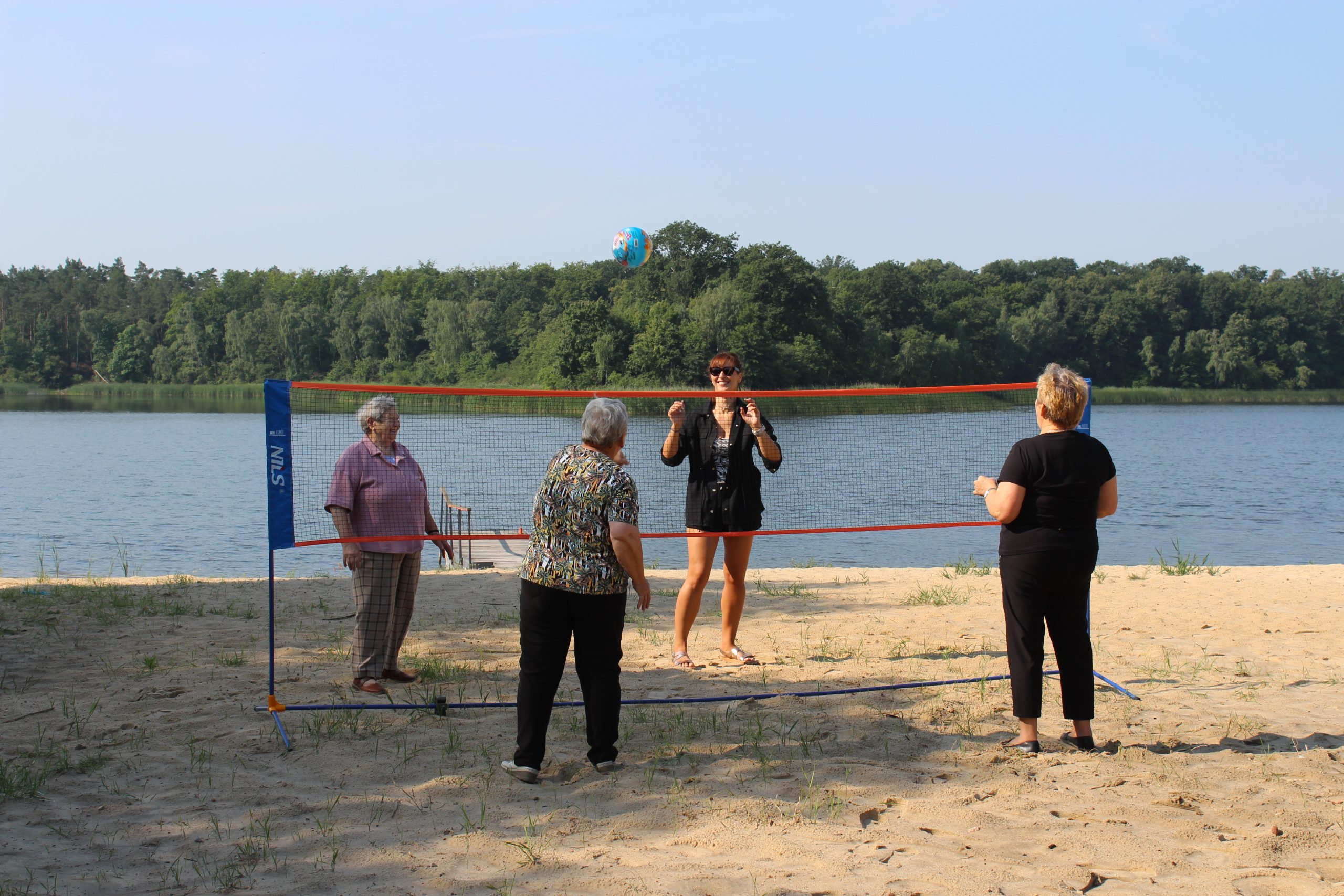 1) Zdjęcie wykonane nad Jeziorem Lubstowskim. Przedstawia cztery kobiety grające w piłkę siatkową na plaży. W tle woda i las.