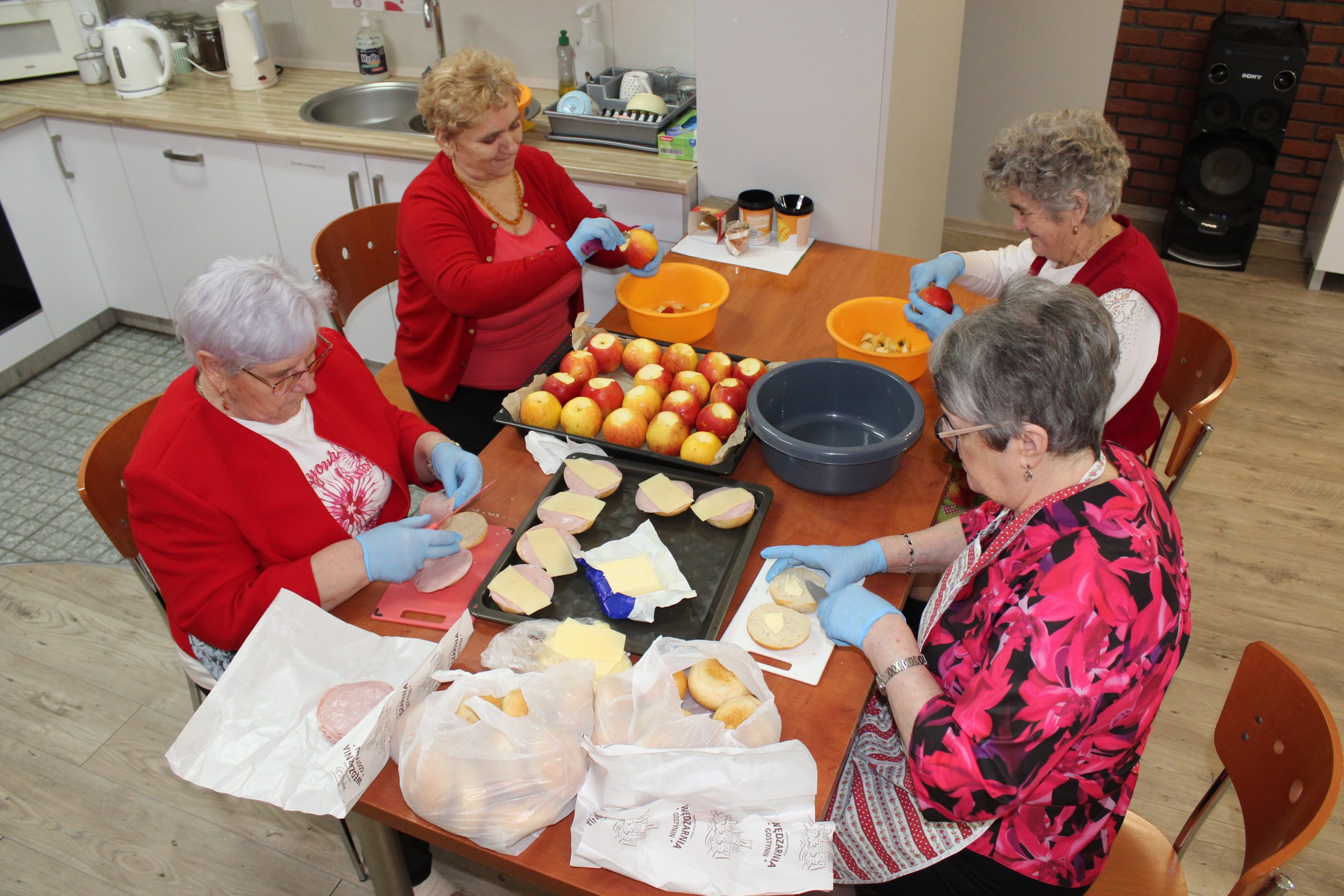 Zdjęcie wykonane w pracowni kulinarnej domu dziennego pobytu. Przedstawia cztery seniorki przygotowujące jabłka i zapiekanki