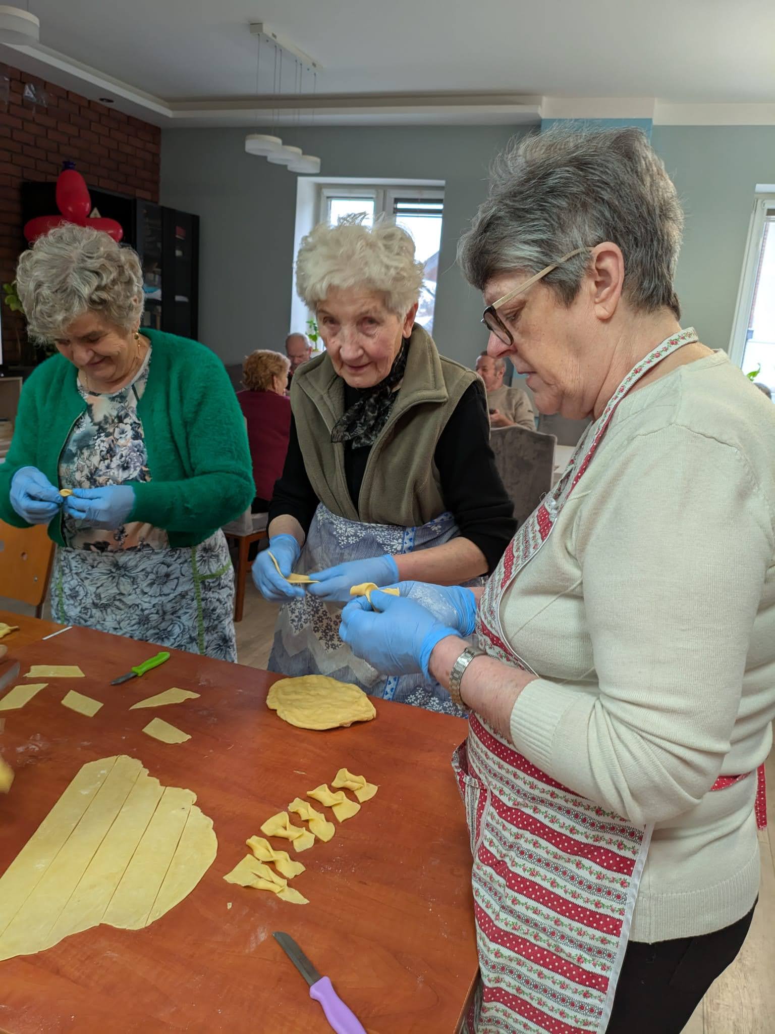 Zdjęcie wykonane w pracowni kulinarnej Domu Dziennego Pobytu w Sompolnie. Przedstawia trzy uczestniczki robiące faworki.