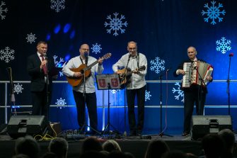 1) Fotografia wykonana w Zespole Szkół Ekonomiczno – Usługowych w Żychlinie. Przedstawia scenę na której stoją przedstawiciele gminy Sompolno: burmistrz Roman Bednarek (wokal), radny Andrzej Prętnicki (gitara), dyrektor MGOPS – Tomasz Majewski (gitara) oraz przedstawiciel seniorów z DDP – Zygmunt Teodorczyk (akordeon). Za nimi rozciąga się niebieska kotara z białymi śnieżynkami.