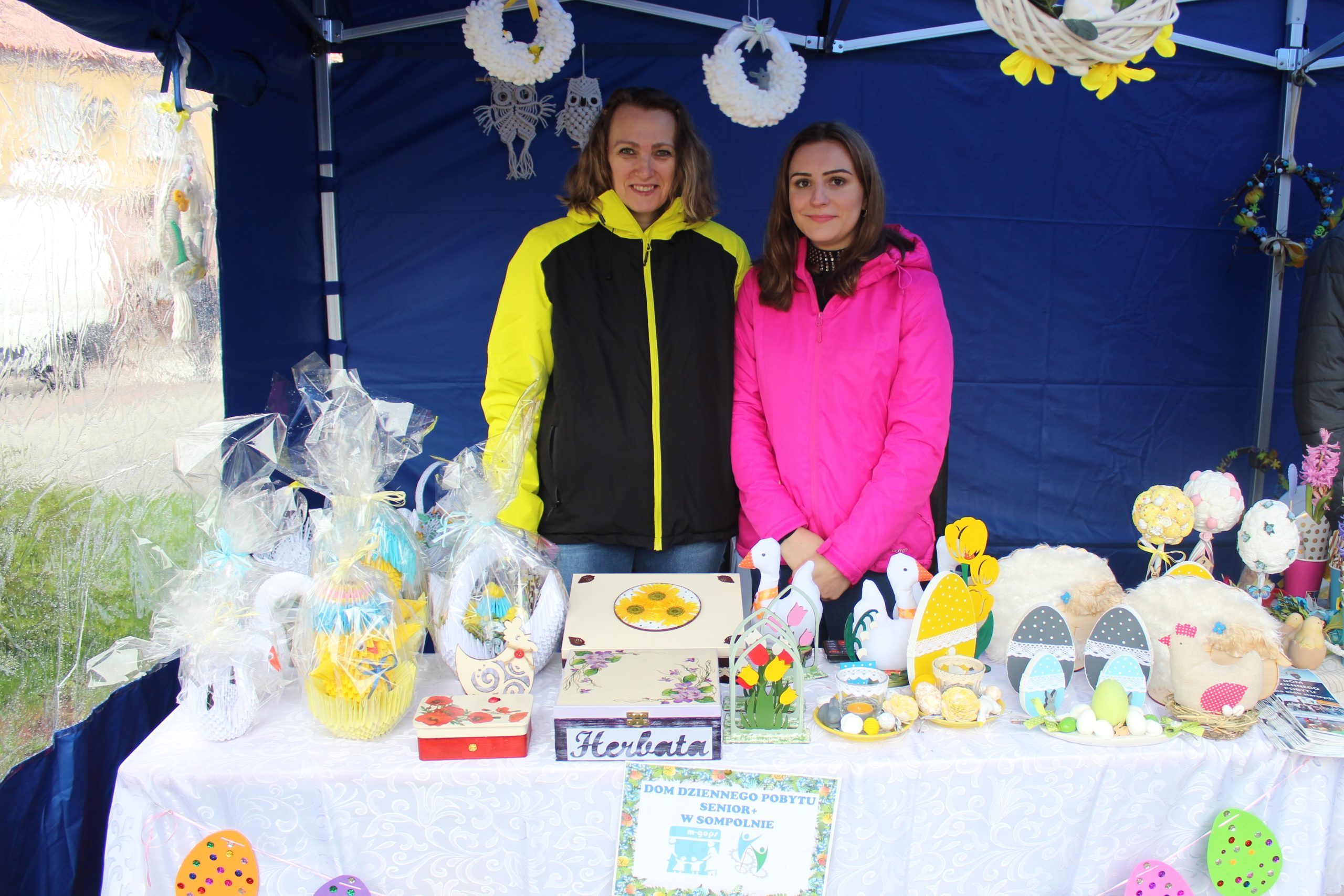 1) Zdjęcie wykonane podczas Sompoleńskiego Kiermaszu Wielkanocnego. Przedstawia stoisko Domu Dziennego Pobytu Senior+ w Sompolnie. W tle dwie kobiety – pracownice DDP.