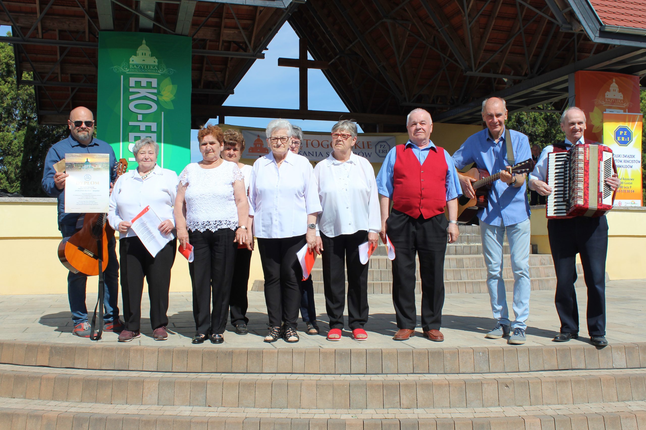 1) Fotografia wykonana na plenerowej scenie w Świętej Górze. Na zdjęciu seniorzy z grupy wokalno – śpiewaczej Wiecznie Młodzi wraz z instruktorem oraz dyrektorem Miejsko – Gminnego Ośrodka Pomocy Społecznej w Sompolnie.