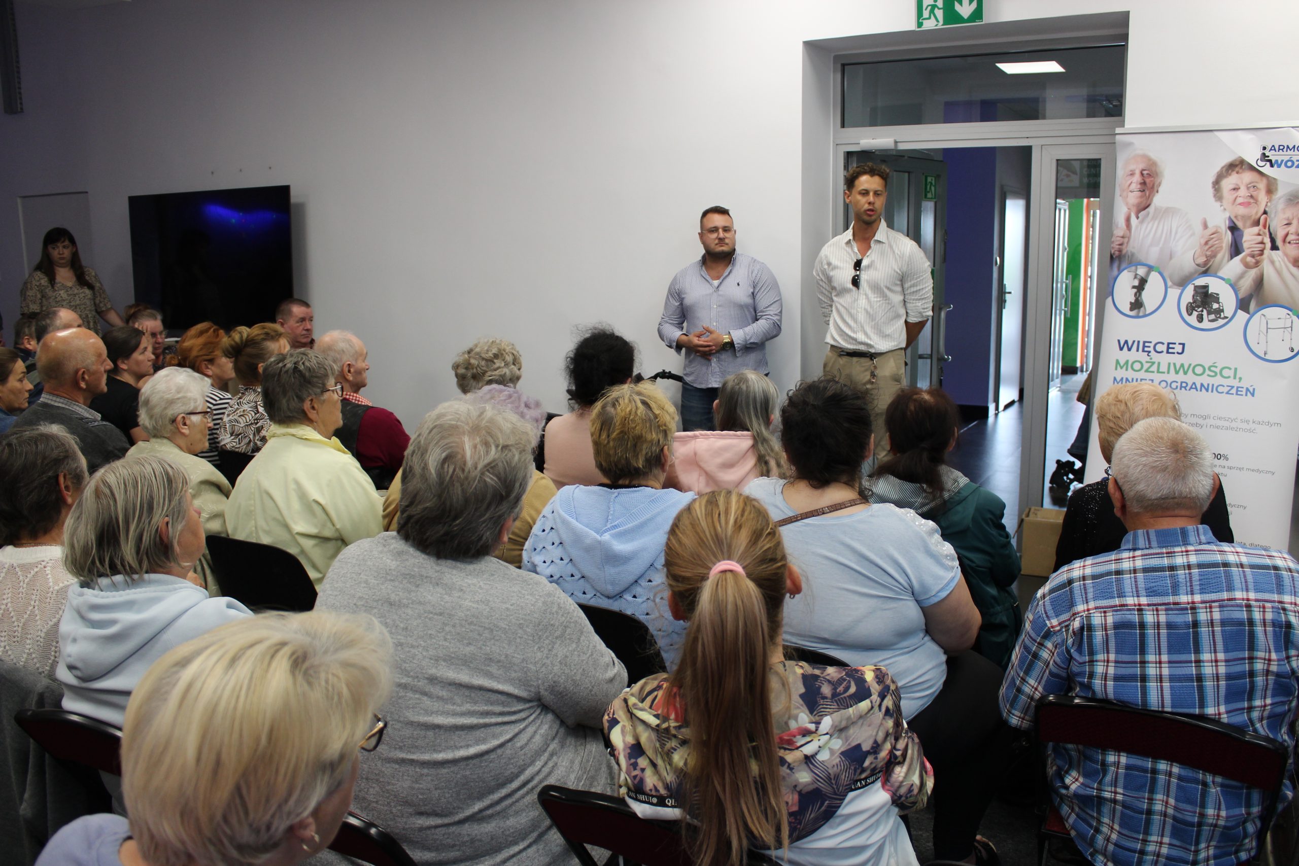 2) Na zdjęciu przedstawiciele Fundacji „Złote lata” opowiadający o celach swojej działalności. Po prawej stronie baner z napisem „Więcej możliwości – mniej ograniczeń”, przed nimi grono osób siedzących na krzesłach.