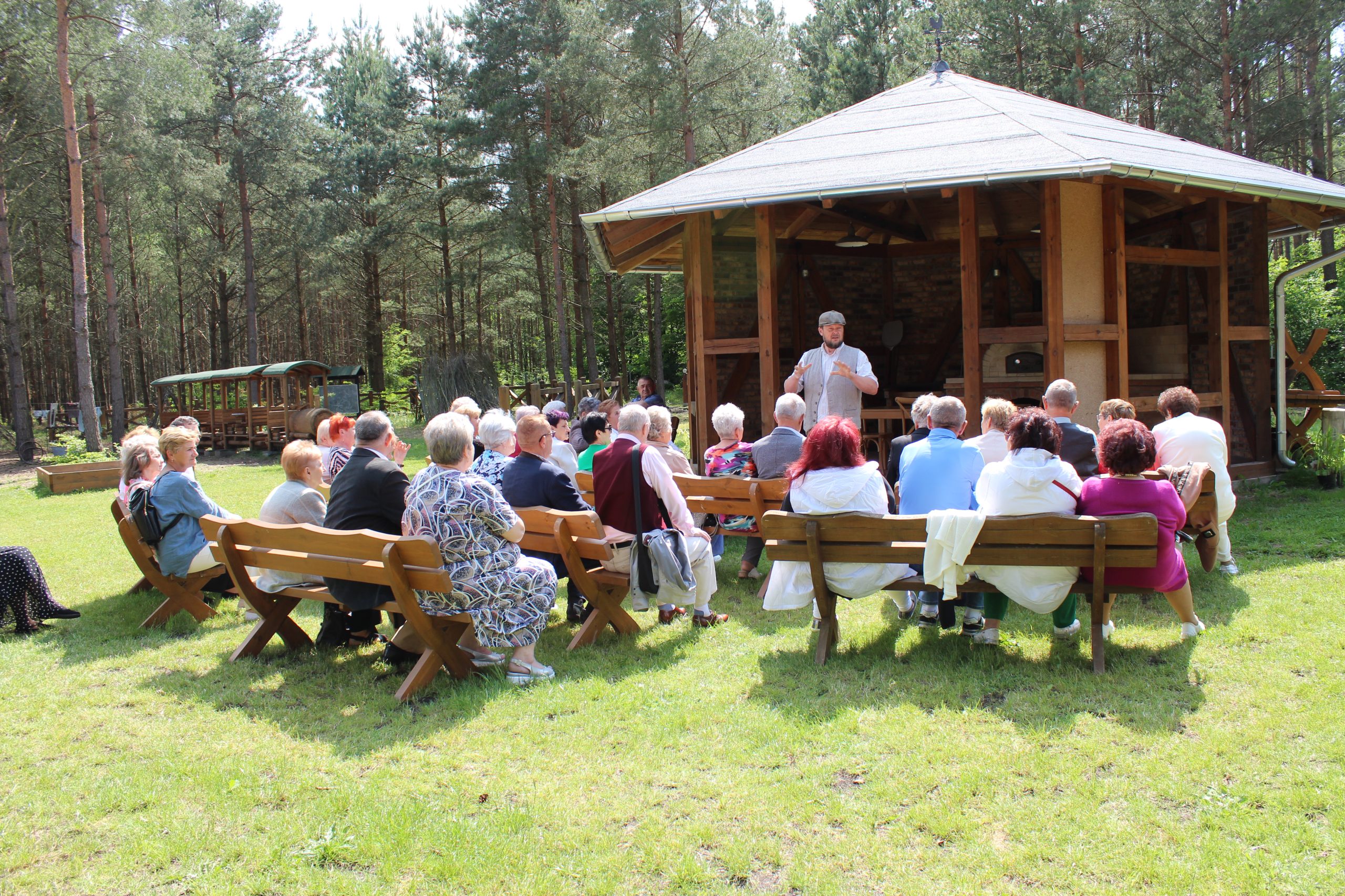 2) Fotografia przedstawia przewodnika, stojącego na tle lasu oraz chaty grillowej, opowiadającego o historii powstania górniczej wioski. Przed przewodnikiem grupa seniorów siedząca na ławkach.