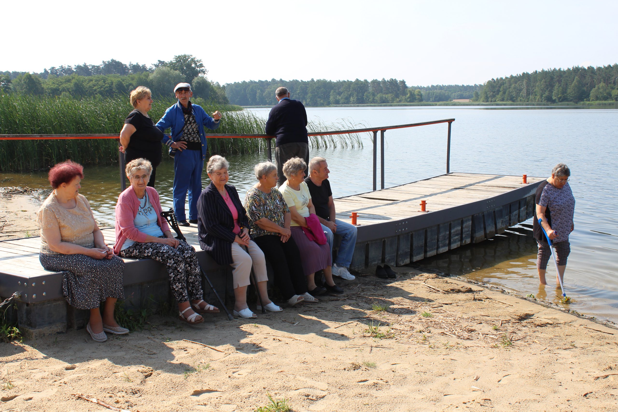 2) Na zdjęciu seniorzy siedzący na pomoście nad jeziorem lubstowskim. Jedna z seniorek spaceruje na brzegu jeziora podpierając się kulą.
