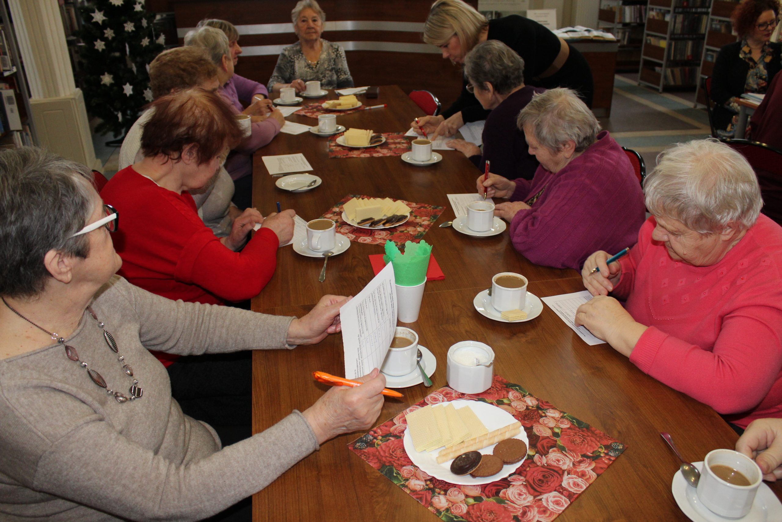 Na zdjęciu widać grupę seniorek siedzących przy stole i rozwiązujących test na kartkach.