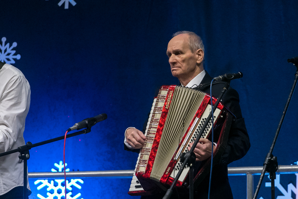 2) Zdjęcie przedstawia starszego mężczyznę grającego na akordeonie. Przed nim mikrofony, za nim niebieskie tło.