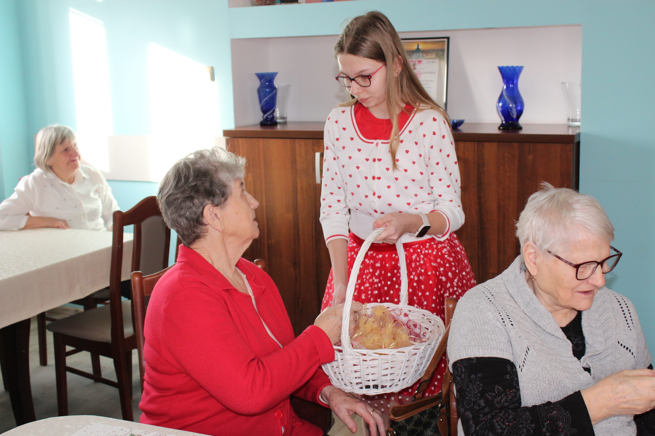 3. Na zdjęciu znajduje się dziewczynka , która trzyma koszyk i wręcza seniorce upominek