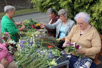3) Na fotografii pięć pań siedzących przy stole podczas wyplatania wianków. Na stole stos kwiatów i ziół.