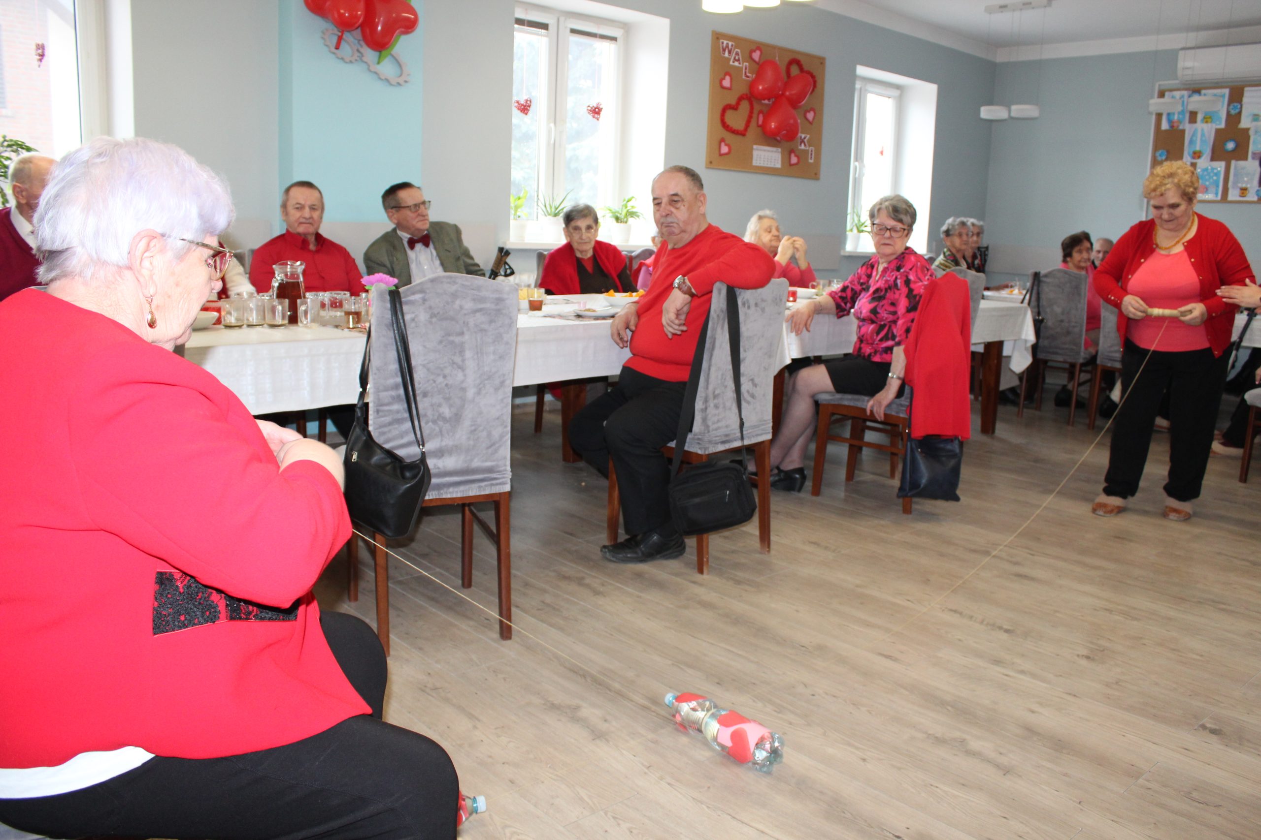 Zdjęcie przedstawia dwie seniorki uczestniczące w zabawie z butelkami. W tle seniorzy siedzący przy stole