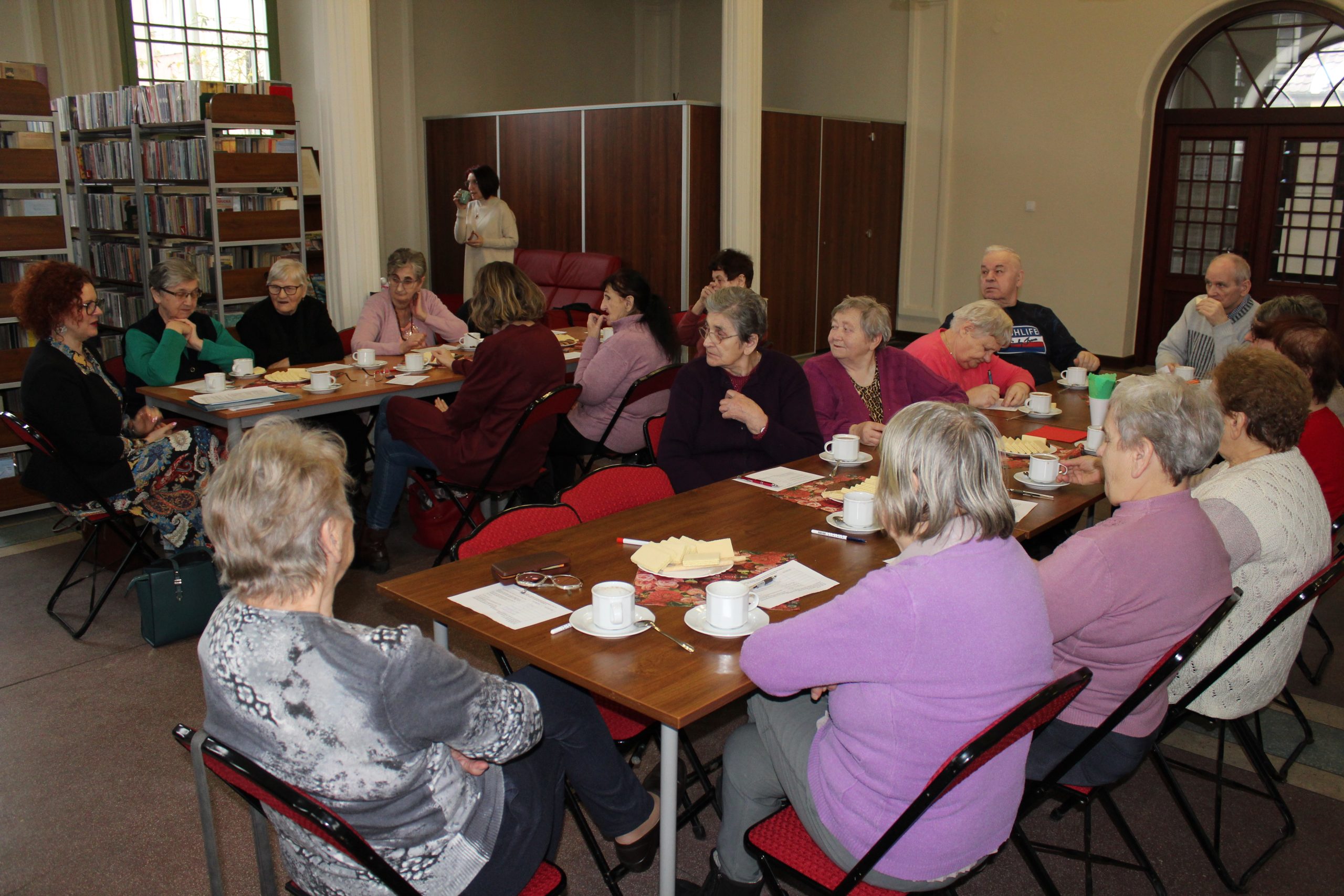 Na fotografii widać seniorów siedzących przy dwóch stołach. Wzrok seniorów skierowany jest na kobietę, która prowadzi wykład o zdrowym żywieniu. W tle regały z książkami.