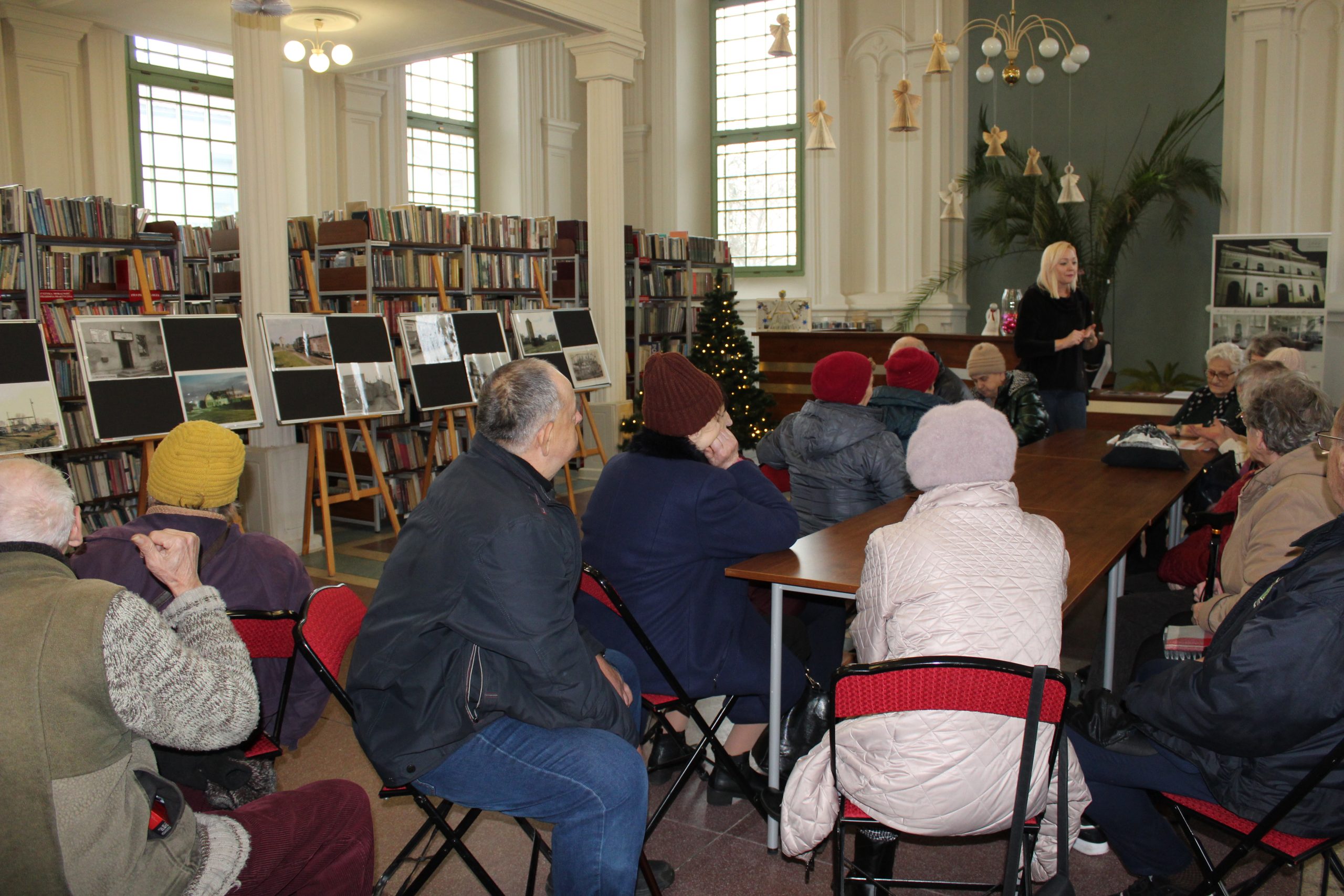4) Zdjęcie wykonane w bibliotece. Przedstawia dyrektorkę biblioteki i seniorów siedzących przy stole. Z lewej strony wystawa fotografii sompoleńskiej kolei wąskotorowej,