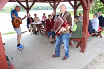 5) Zdjęcie przedstawia mężczyzn grających na akordeonie i gitarze. W tle seniorzy siedzący na ławkach przy stołach pod altaną grillową.