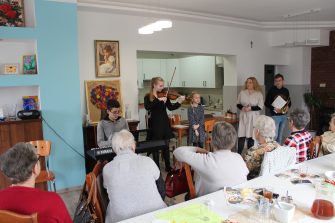 5) Na zdjęciu wszyscy uczniowie wraz z nauczycielką, którzy przyszli do DDP zaprezentować koncert wiosenny, oraz seniorzy siedzący przy stole wsłuchani w grę na instrumentach.