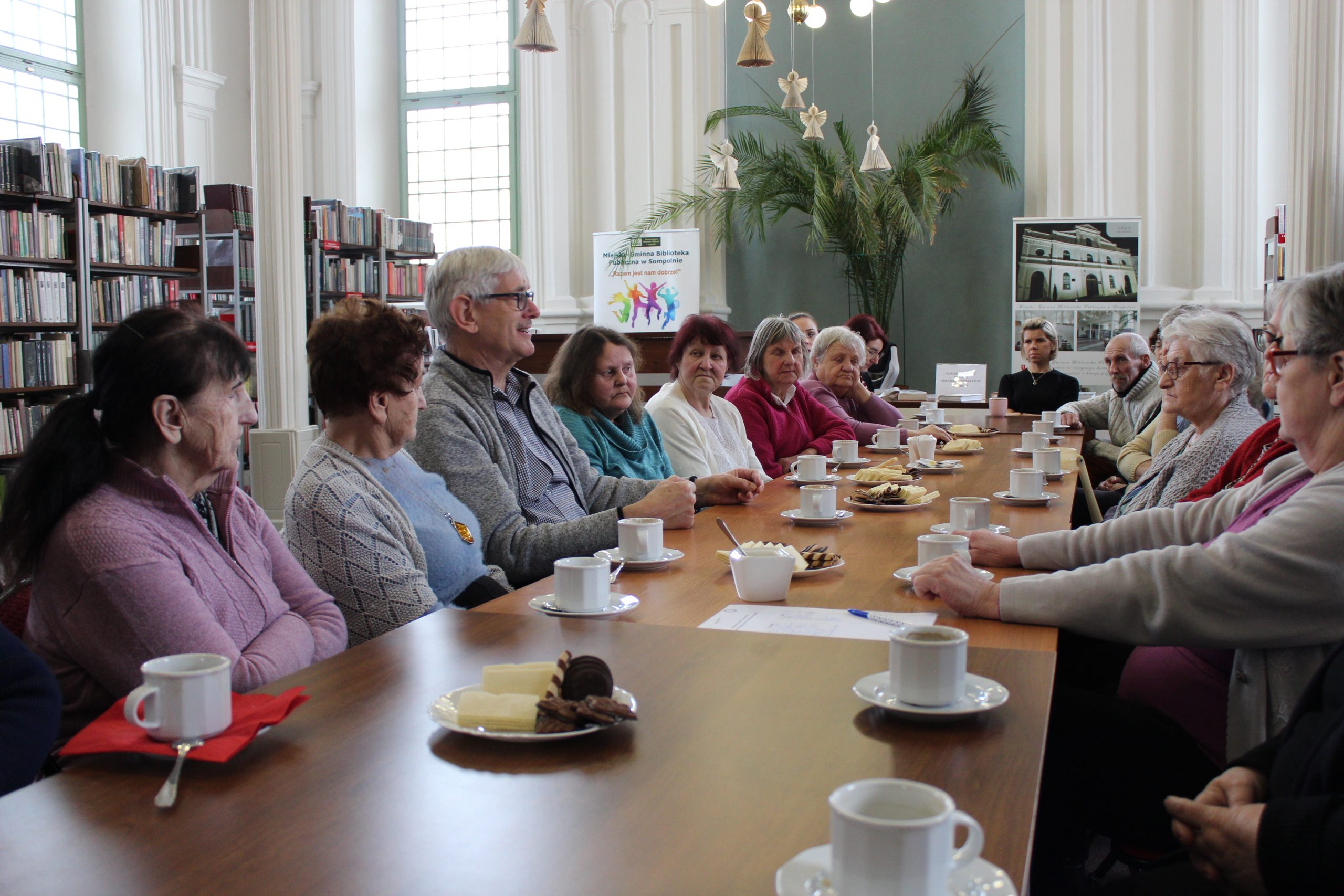 6) Fotografia przedstawia grupę seniorów siedzącą przy wspólnym stole wraz z wykładowcą w bibliotece.