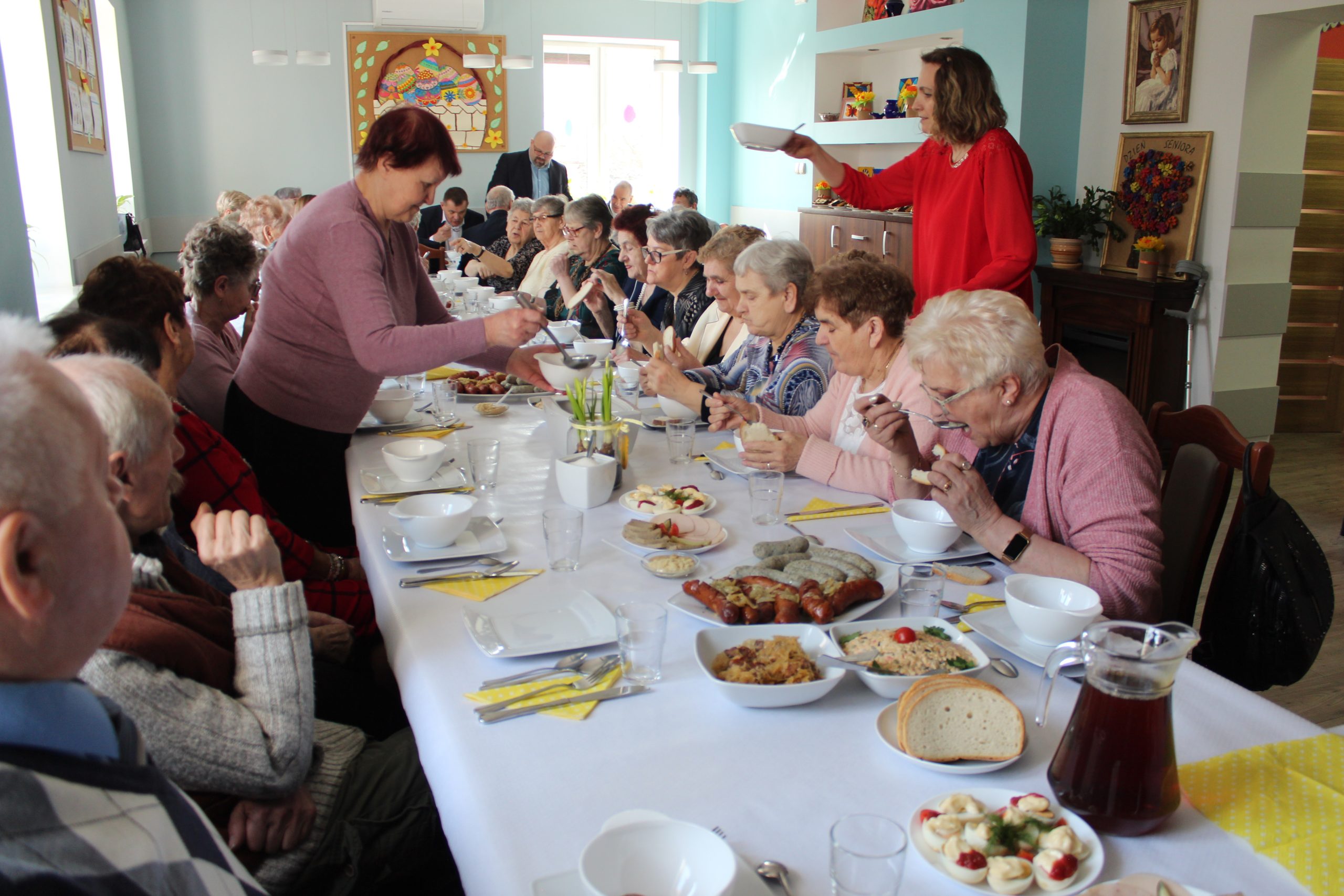 6 Zdjęcie przedstawia seniorów siedzących przy stole i spożywających śniadanie wielkanocne. Po prawej stronie jedna z pań nalewa żurek, a po lewej stronie opiekunka osób starszych donosi miskę z jedzeniem.