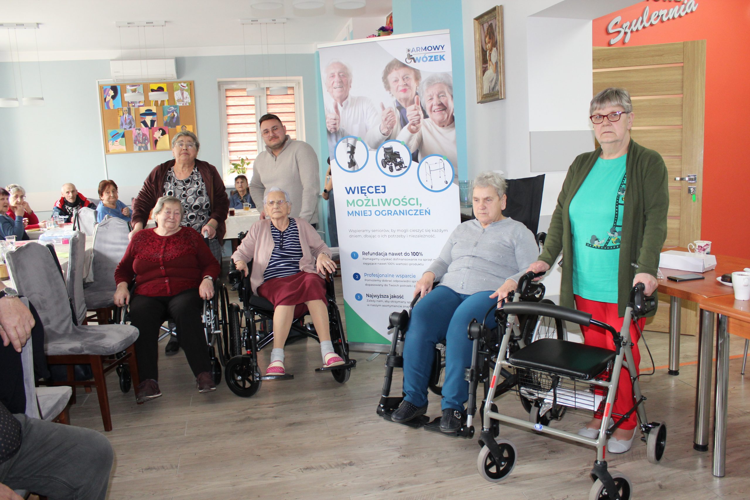 Zdjęcie prezentuje uczestniczki ddp siedzące na wózkach inwalidzkich, panią z balkonikiem oraz konsultanta medycznego zebranych wokół banneru – darmowy wózek.
