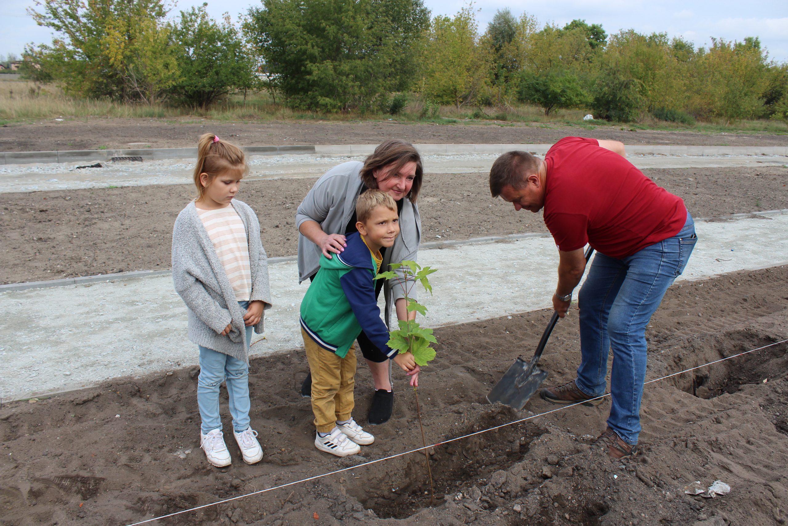 9) Fotografia prezentuje kobietę z dziećmi oraz burmistrza, który sadzi drzewo.