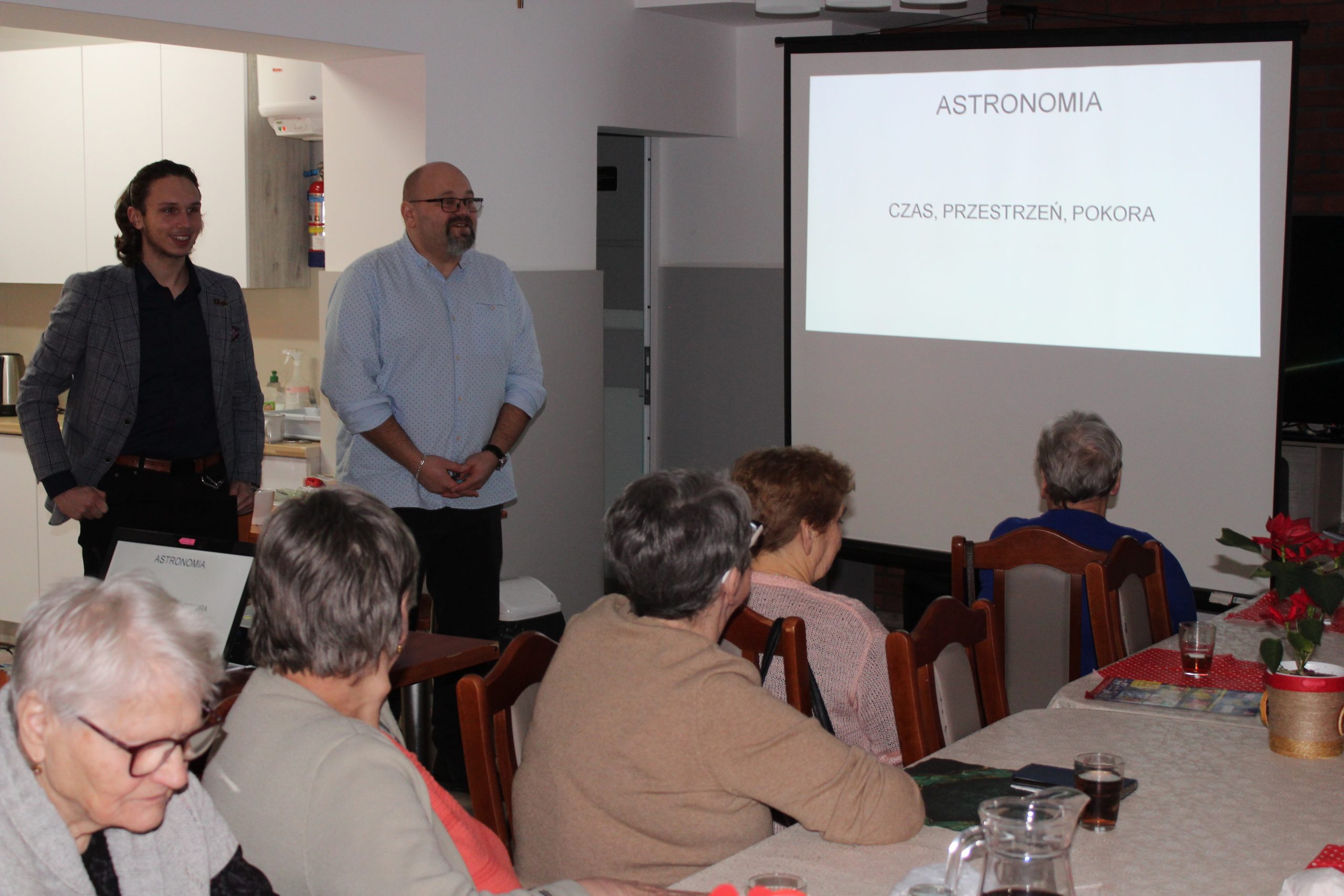 1) Zdjęcie wykonane na sali głównej Domu Dziennego Pobytu dla Osób Starszych w Sompolnie. Widać na nim dyrektora MGOPS w Sompolnie oraz Pana Mikołaja Błaszczyka – ucznia liceum w Sompolnie. Po prawej stronie tytuł prezentacji – ASTRONOMIA CZAS PRZESTRZEŃ POKORA. Seniorzy siedzący na krzesłach przy stole.