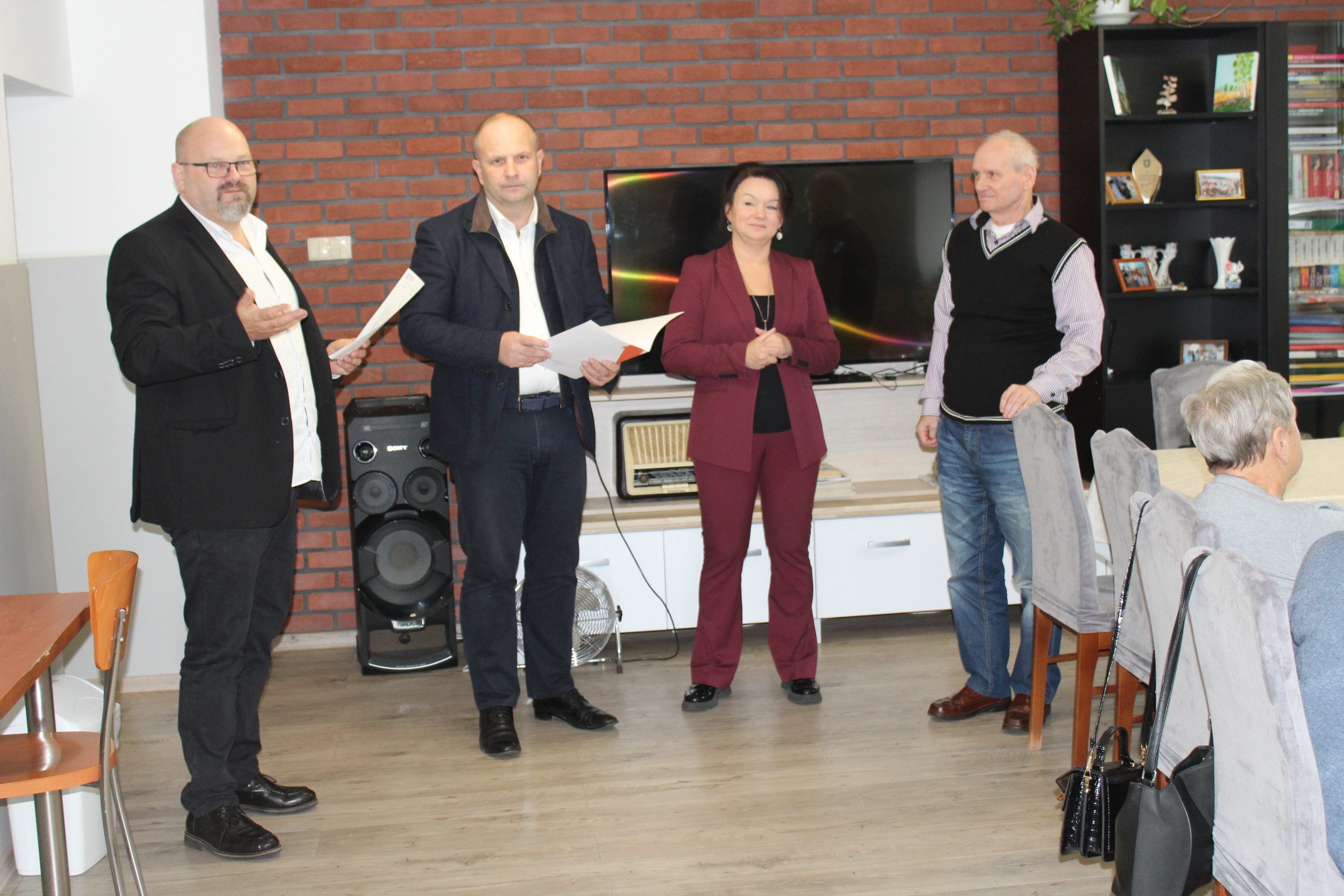 1) Zdjęcie wykonane w sali głównej Domu Dziennego Pobytu dla Osób Starszych w Sompolnie. Przedstawia od lewej: pana Tomasza Majewskiego – dyrektora MGOPS w Sompolnie, pana Waldemara Marciniaka – burmistrza miasta Sompolna, panią Anetę Niestrawską - kierowniczkę administracyjną urzędu miejskiego w Sompolnie oraz pana Zygmunta Teodorczyka – uczestnika DDP