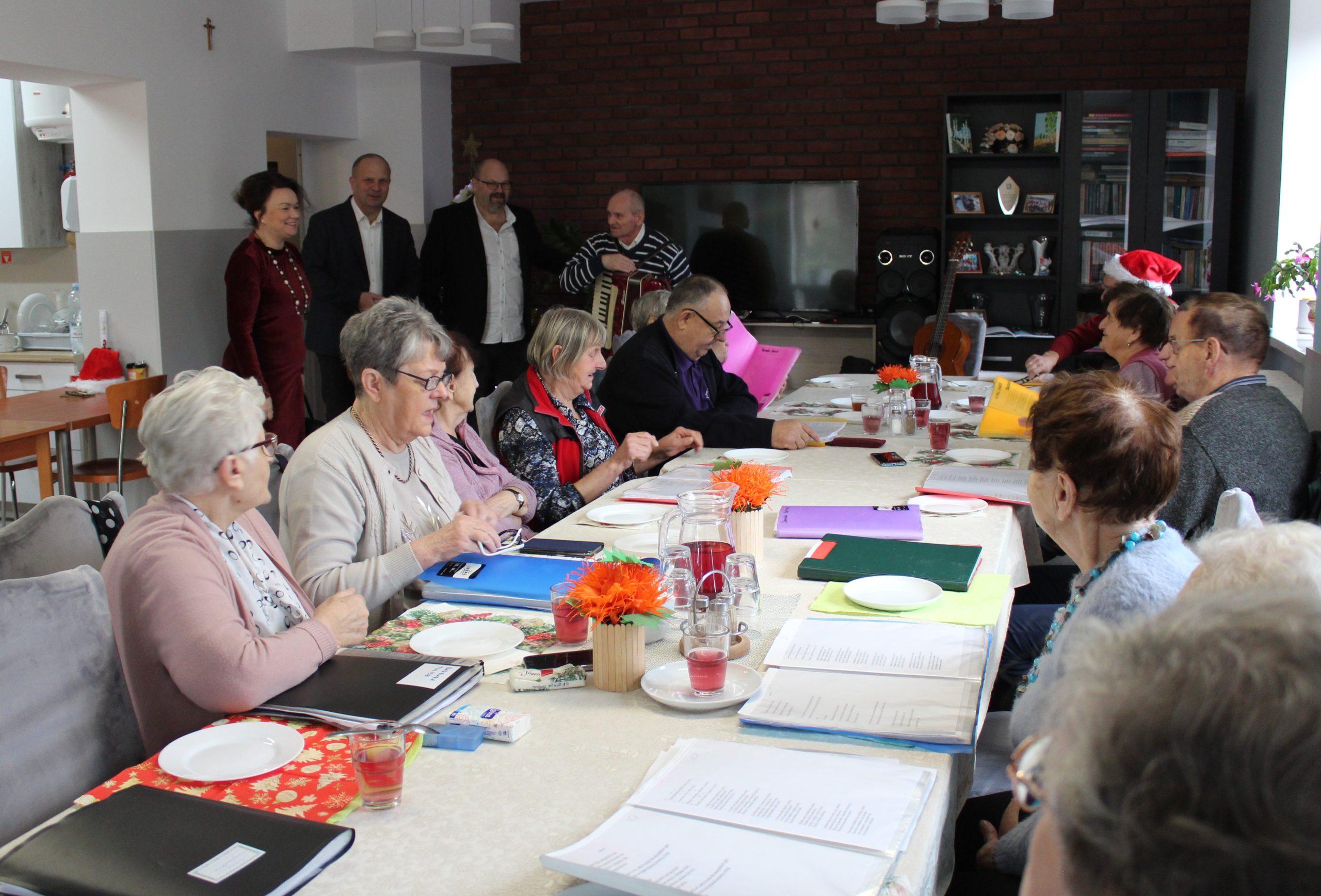 1) Zdjęcie wykonane na sali głównej domu dziennego pobytu. Przedstawia seniorów siedzących przy stole, panią sekretarz, pana burmistrza, dyrektora mgops oraz jednego z uczestników stojącego z akordeonem.