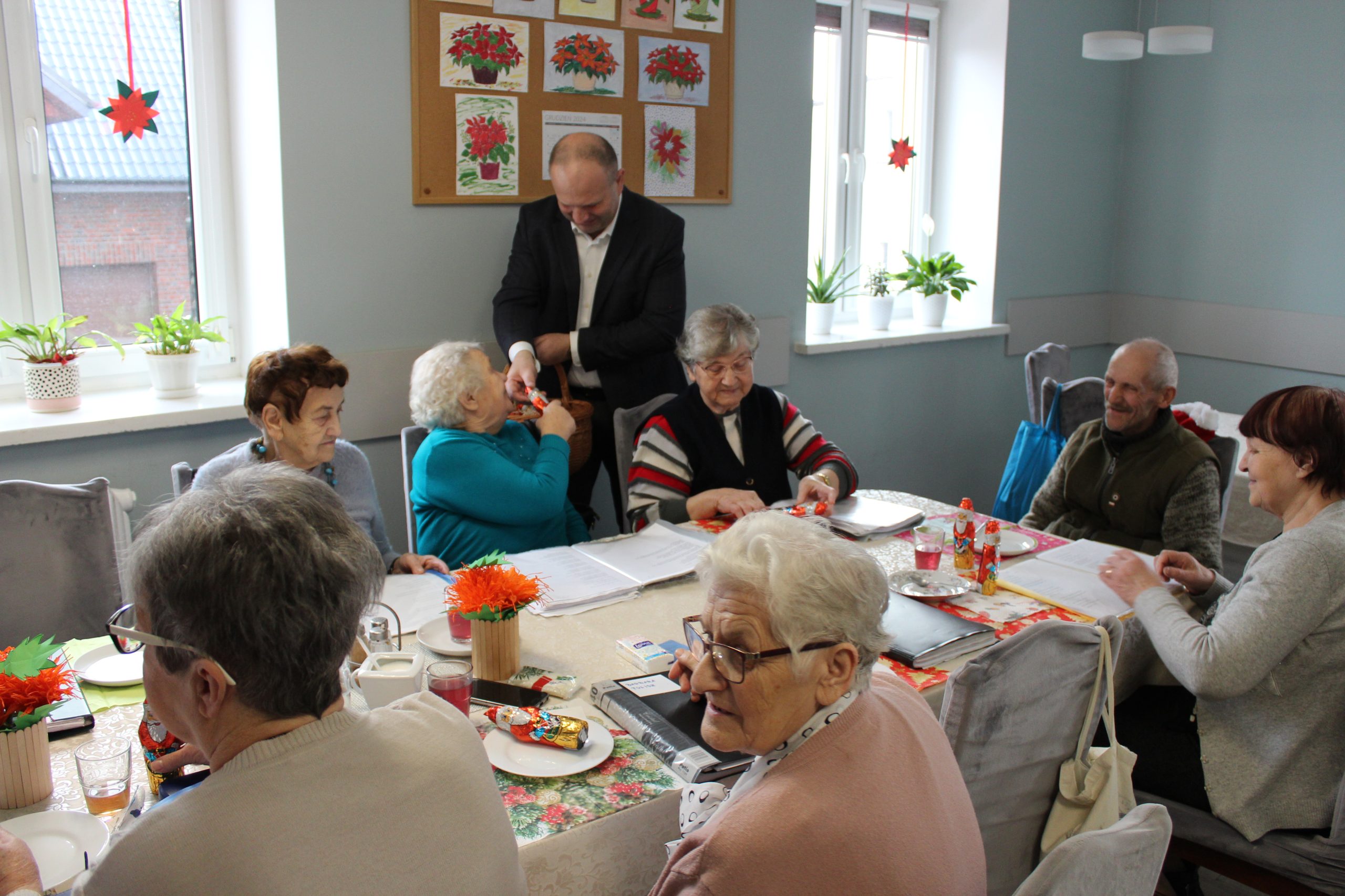 3) Fotografia przedstawia burmistrza rozdającego czekoladowe mikołaje seniorom siedzącym przy stole.