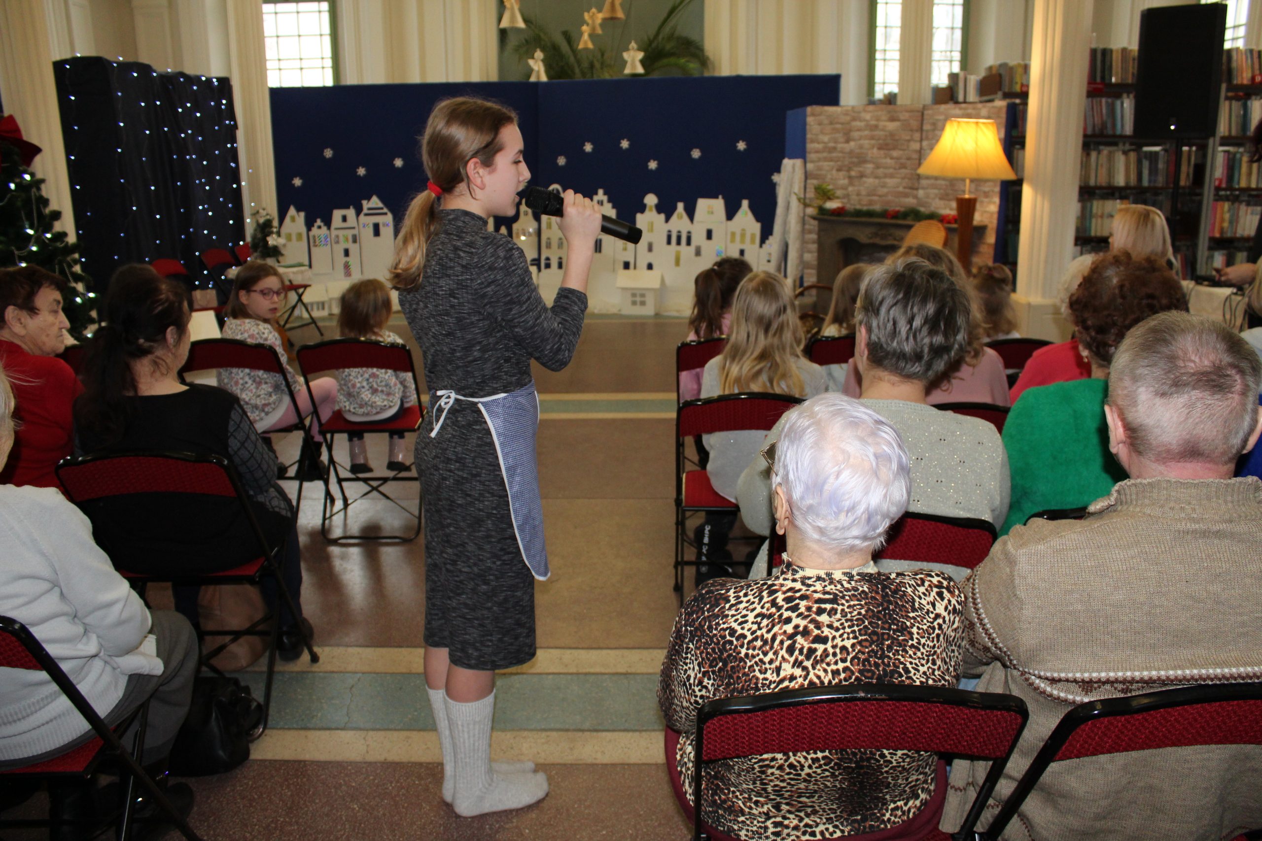 1) Zdjęcie wykonane w miejsko – gminnej bibliotece publicznej w Sompolnie. Przedstawia dziewczynę chodzącą wśród publiczności z mikrofonem w dłoni. W tle scenografia do spektaklu „Dziewczynka z zapałkami”.