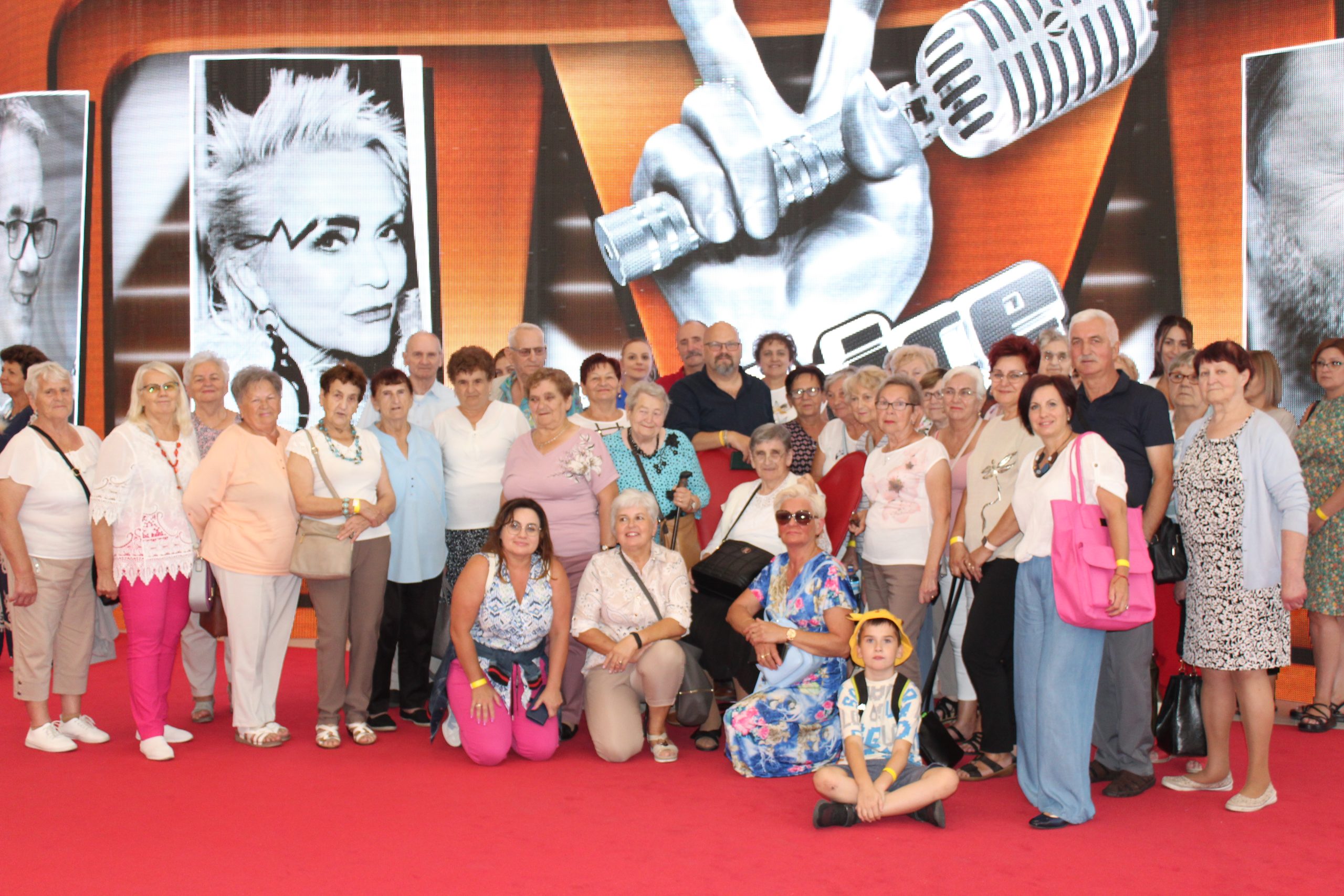 4) Zdjęcie grupowe wykonane w siedzibie telewizji polskiej w Warszawie. Przedstawia grupę seniorów z Sompolna i okolic wraz z opiekunami pozującą na tle plakatu THE VOICE SENIOR.