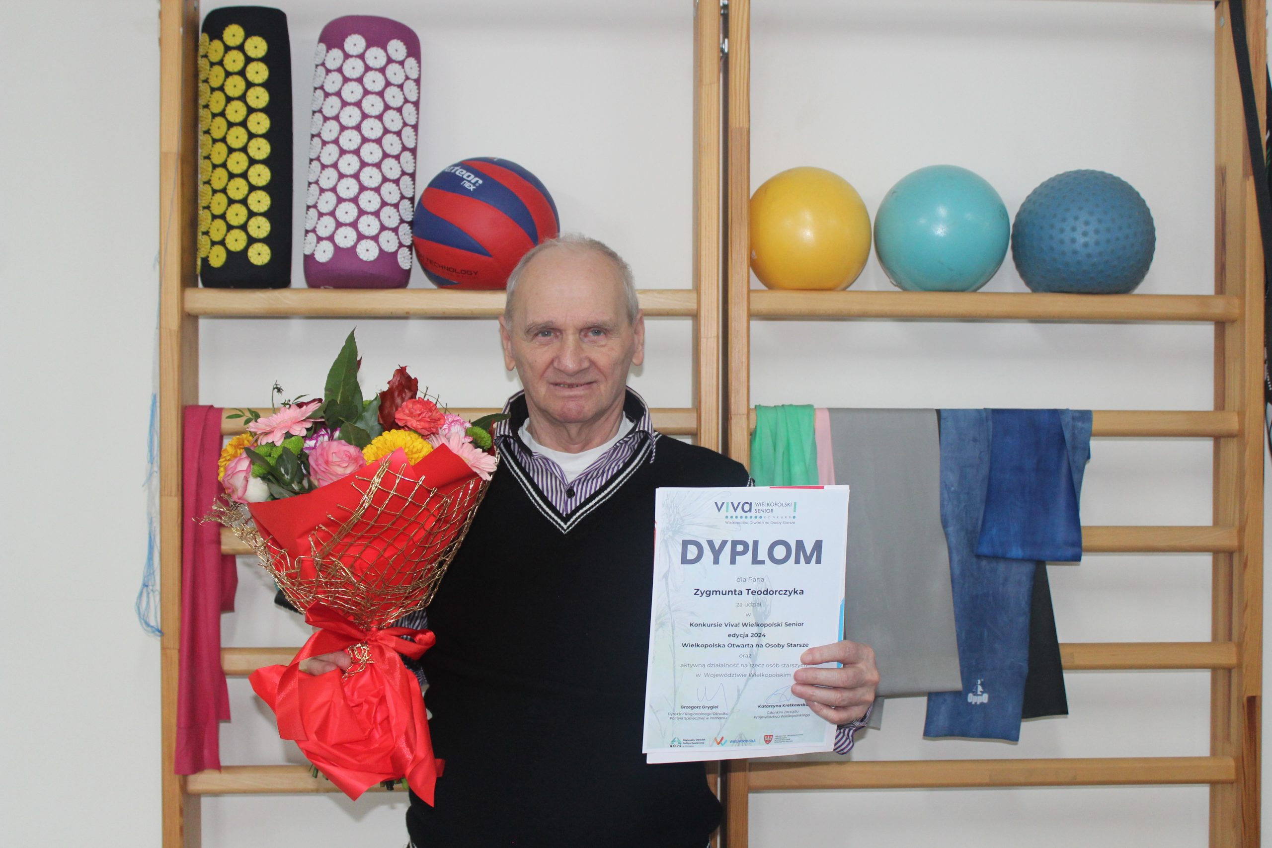 5) Zdjęcie wykonane w sali rehabilitacyjnej przedstawia seniora trzymającego kwiaty i dyplom.