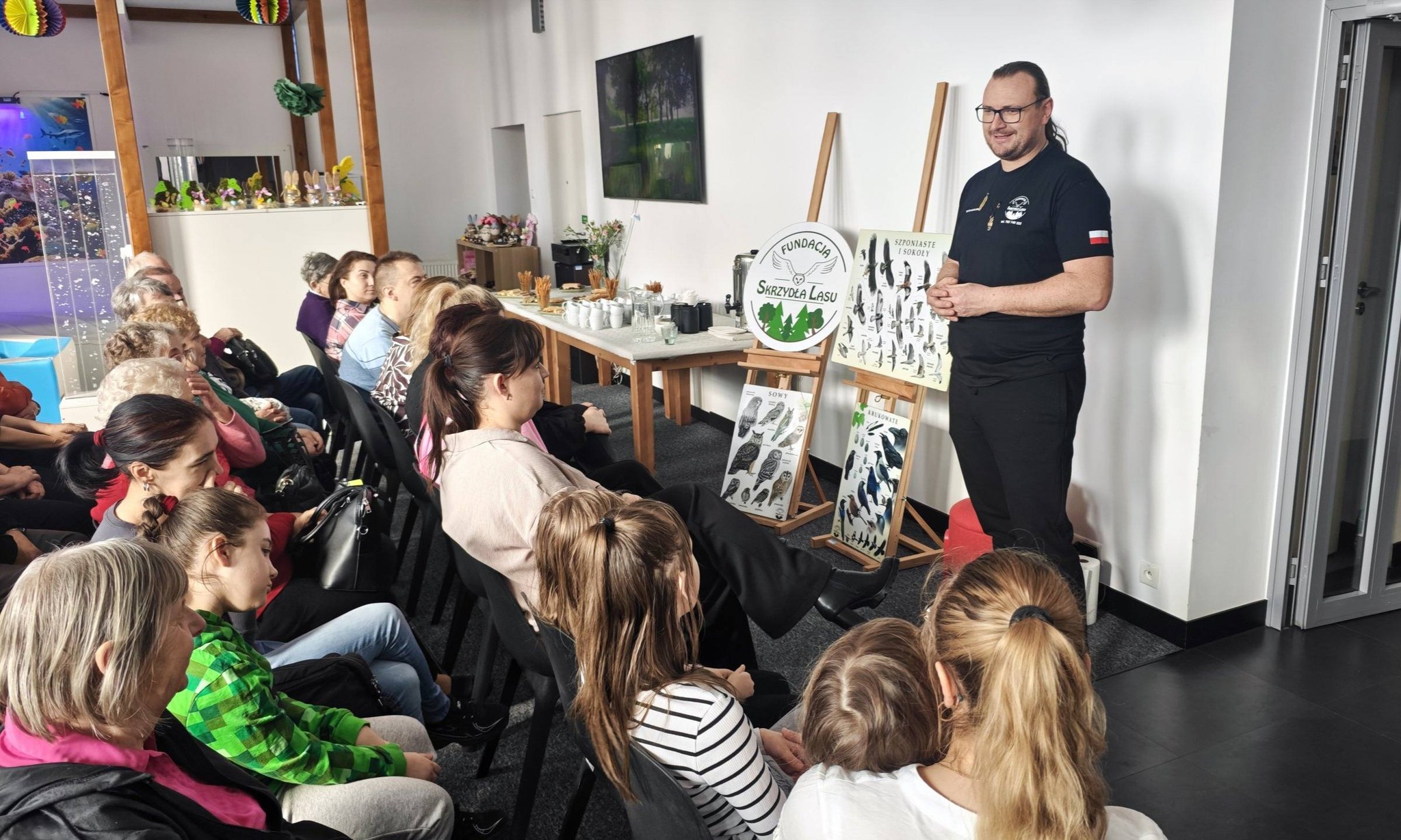 1) Zdjęcie wykonane w sali głównej Dziennego Centrum Wsparcia. Na fotografii przedstawiciel Fundacji Skrzydła Lasu wita zebranych na prezentacji.