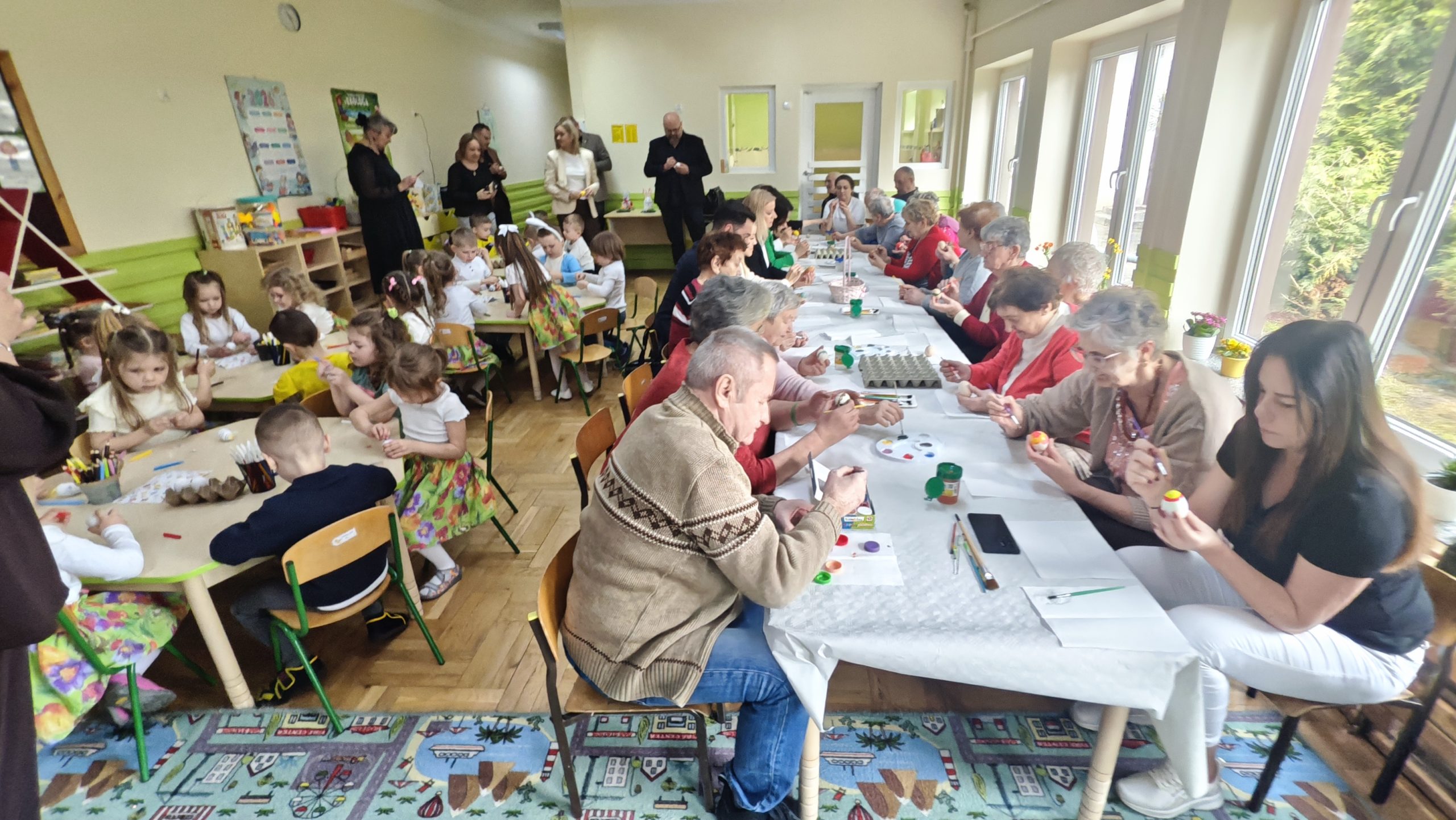 5. Zdjęcie wykonane na na sali przedszkolnej gdzie widać wszystkich zgromadzonych, którzy wspólnie dekorują jajka.