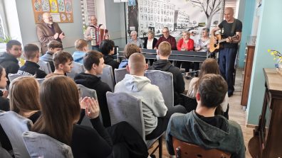 1) Zdjęcie wykonane w sali głównej Domu Dziennego Pobytu. Przedstawia dyrektora MGOPS, instruktora muzykoterapii, seniorów oraz młodzież podczas przedstawienia teatralnego.