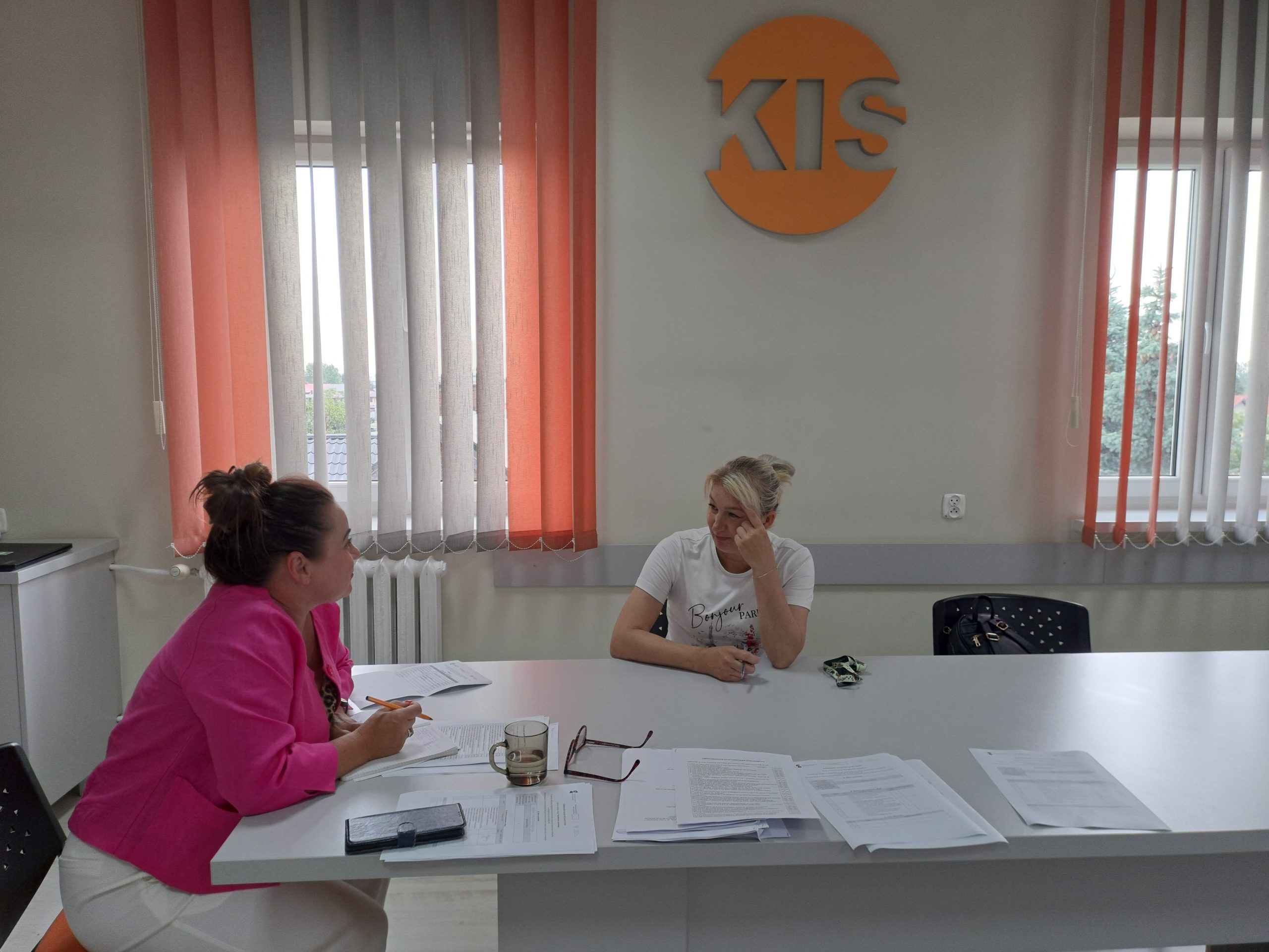 Na zdjęciu widzimy dwie kobiety rozmawiające ze sobą. Jedna z nich to uczestniczka kursu. Druga kobiety trzyma w dłoni długopis, a przed sobą ma plik dokumentów.